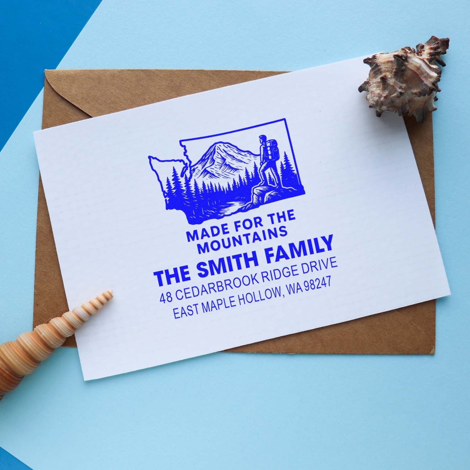 Custom family address stamp with mountain hiker design on white card over brown envelope, styled with seashells on a blue background.