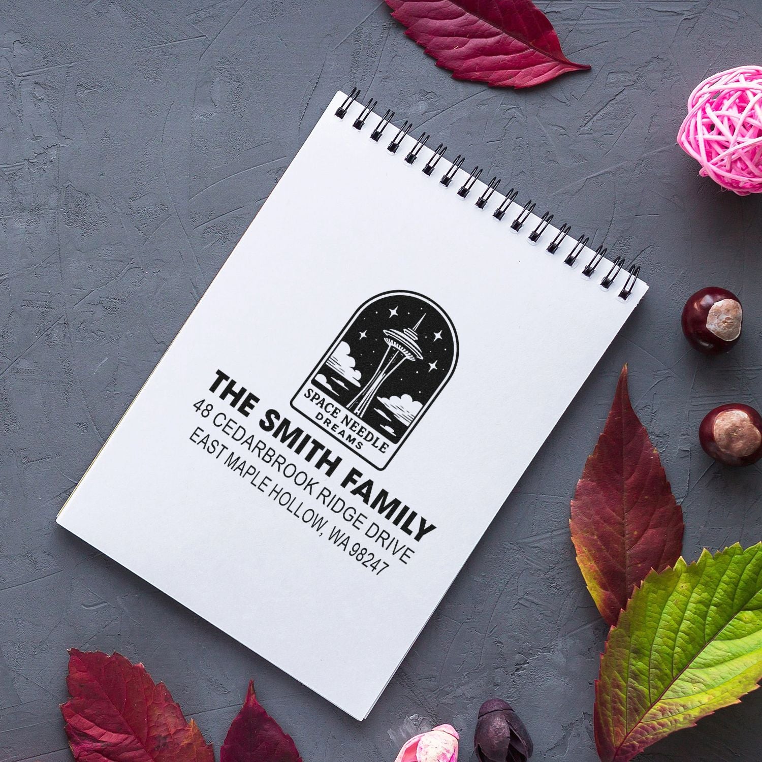 Custom return address stamp for The Smith Family with Space Needle design on a spiral notebook, styled on a gray tabletop with autumn leaves and decor.