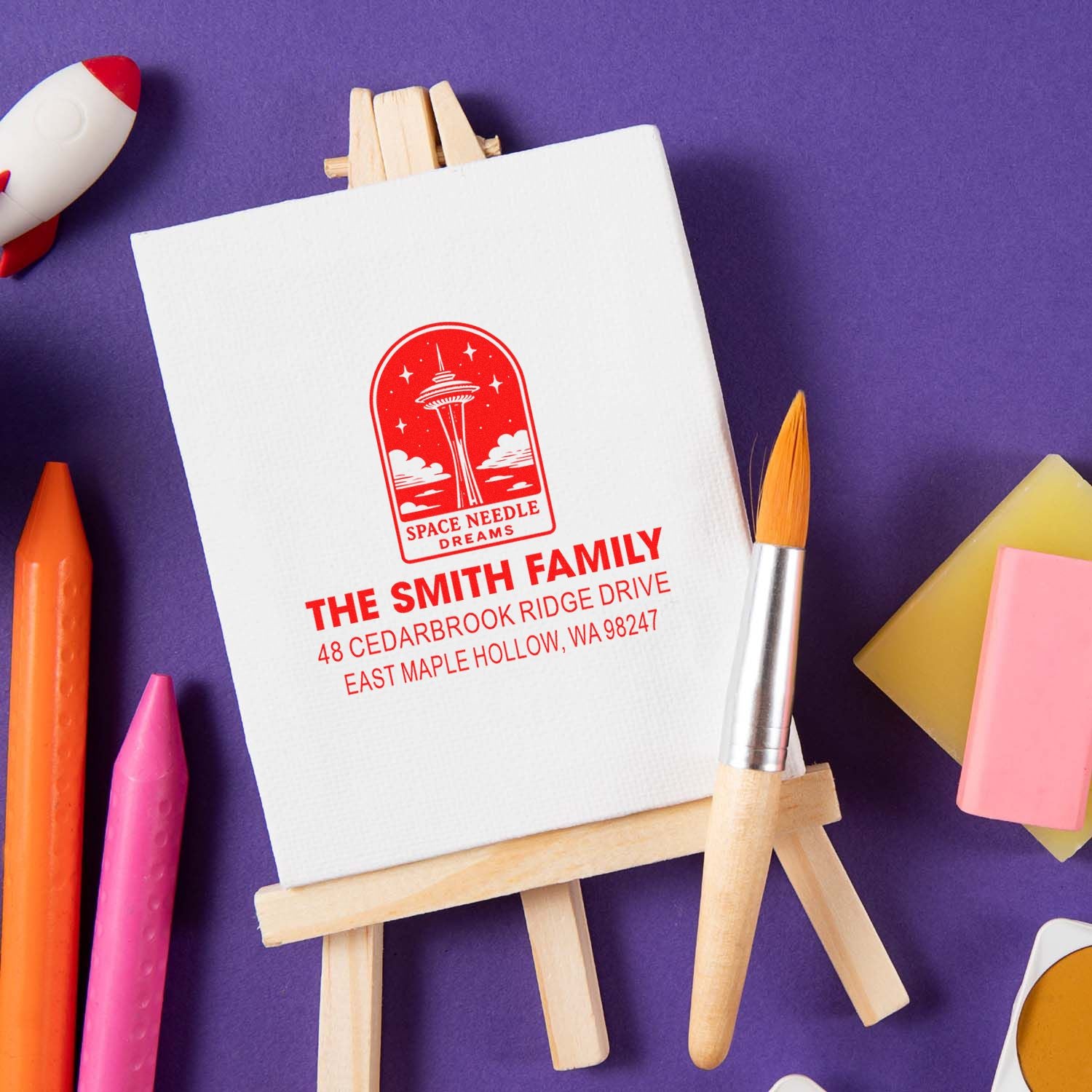 Custom return address stamp with Space Needle Dreams design on an easel, surrounded by art supplies on a purple background.