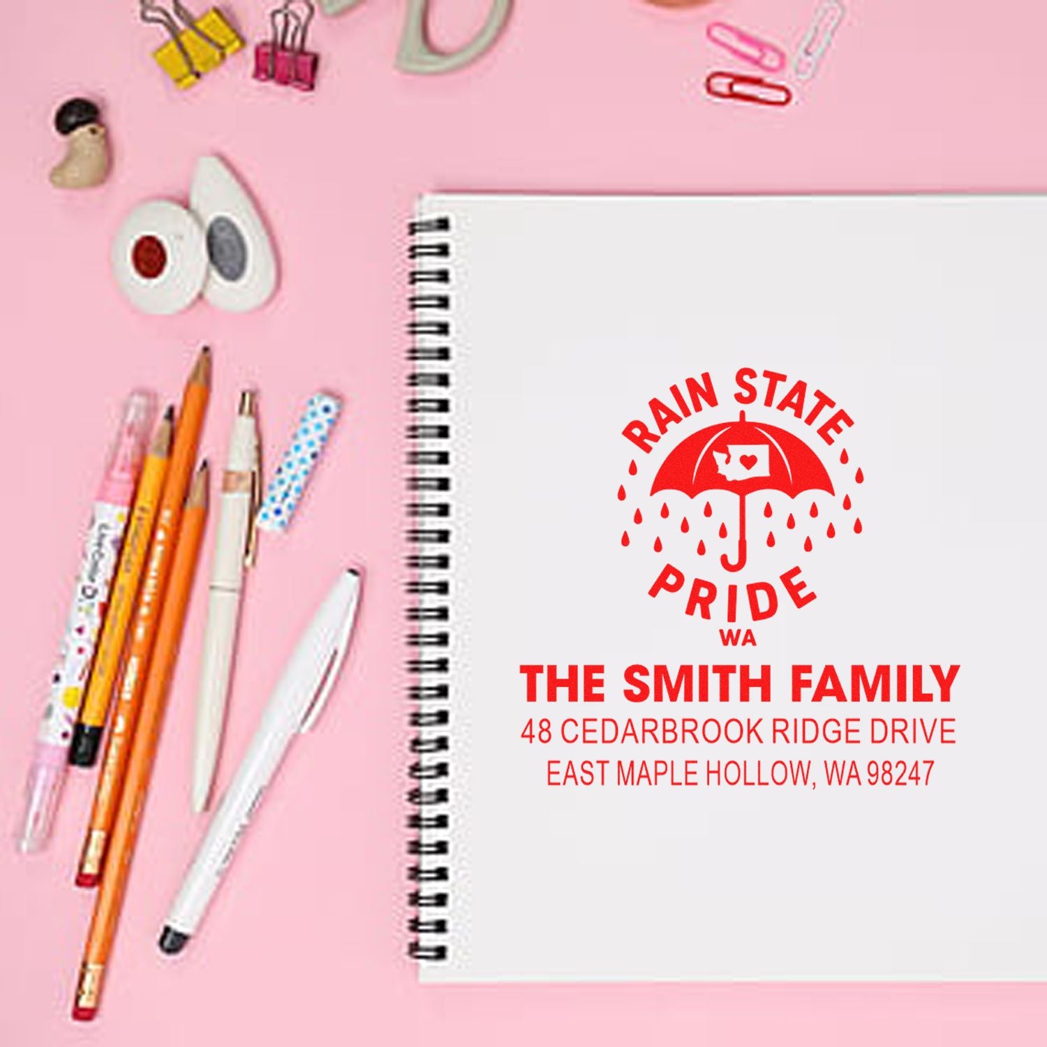 Custom Rain State Pride address stamp on a spiral notebook, styled on a pink desk with pens, binder clips, and stationery.