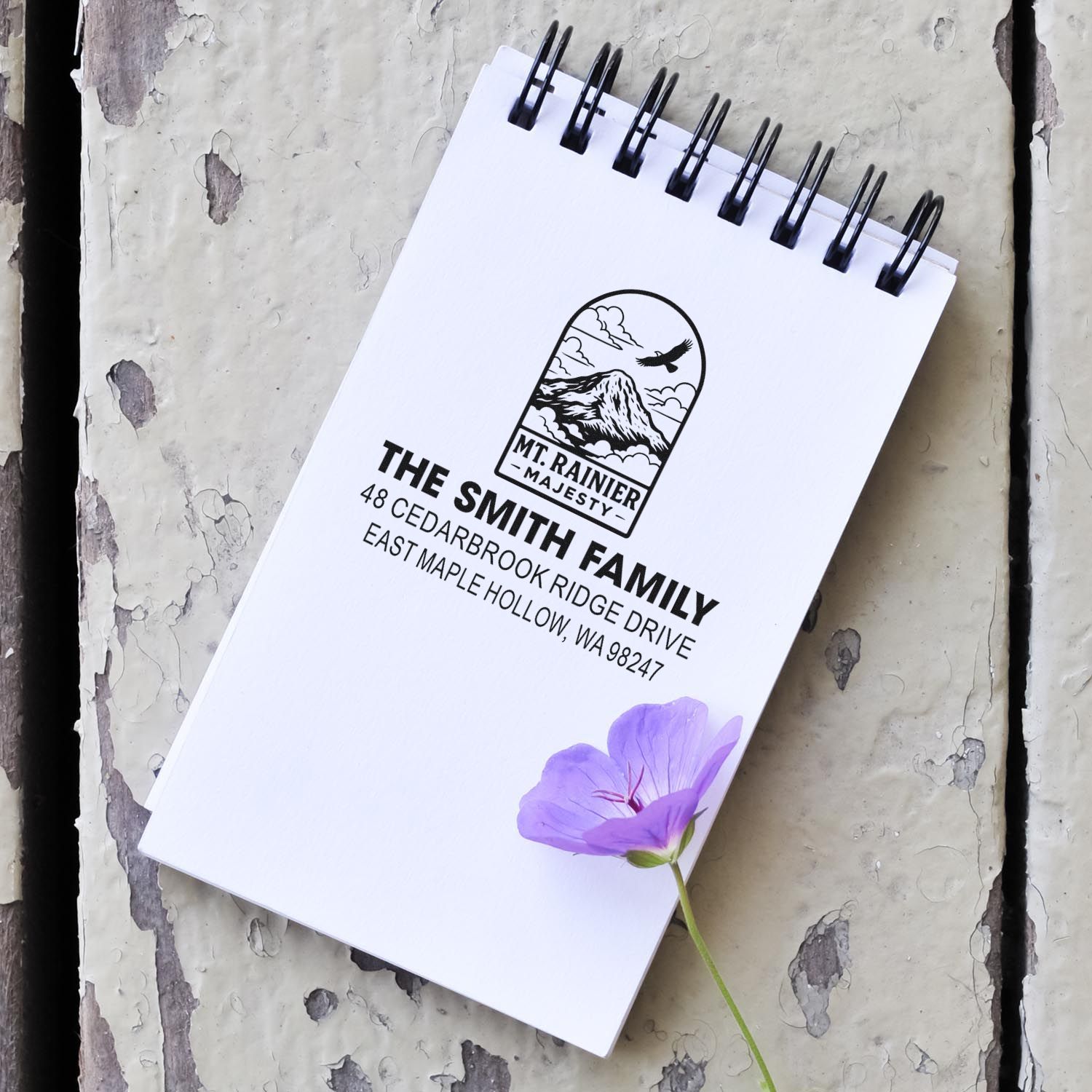 Custom Mount Rainier address stamp on a white spiral notepad, with purple flower, set on weathered painted wood background.