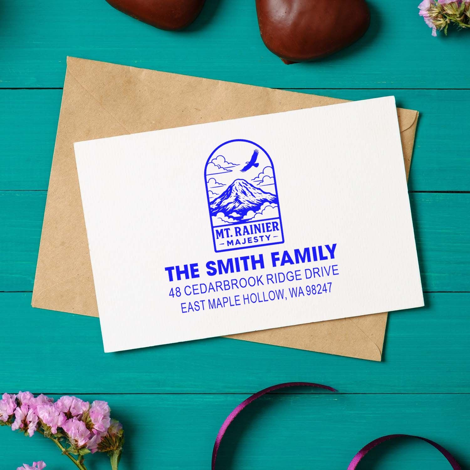 Custom Mt. Rainier return address stamp for The Smith Family on a white card over a kraft envelope, styled on a teal wooden tabletop with flowers.