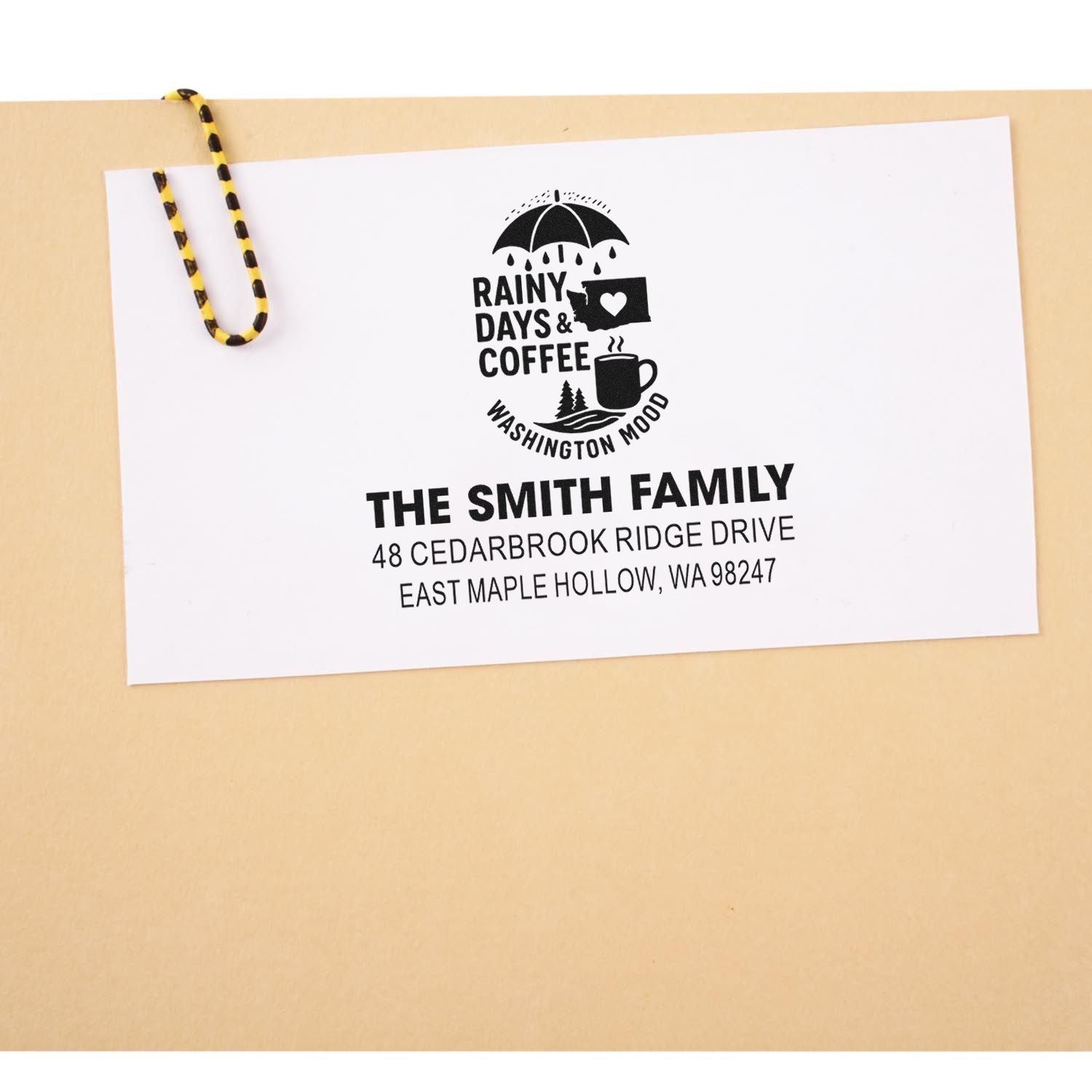 Custom address stamp on white card with rainy days and coffee design, clipped to tan background with yellow-black paperclip.