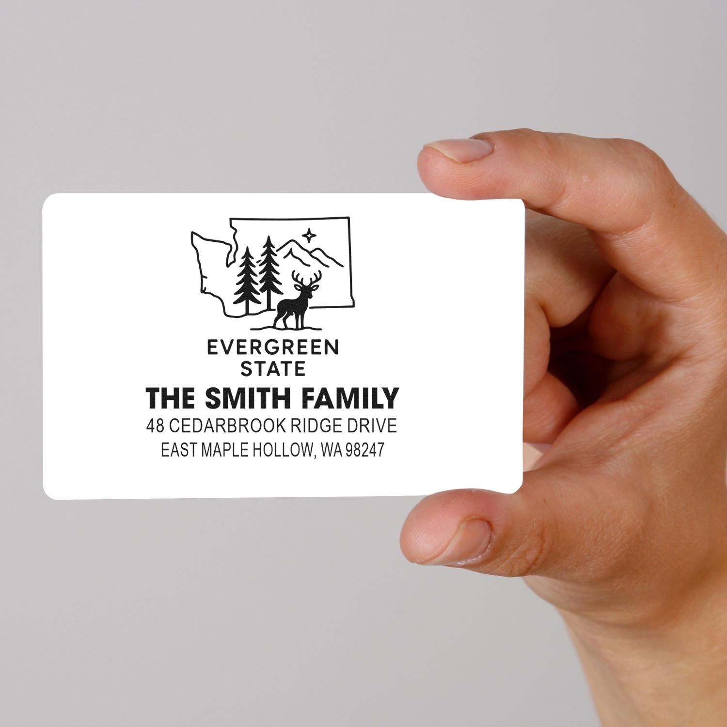Hand holding a personalized return address stamp with Washington state outline, evergreen trees, deer, and The Smith Family address on a gray background.