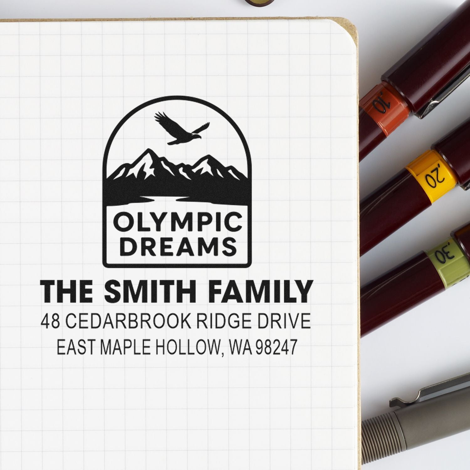 Custom address stamp for The Smith Family featuring Olympic Dreams mountain eagle logo, shown on grid paper beside colorful self-inking stamps.