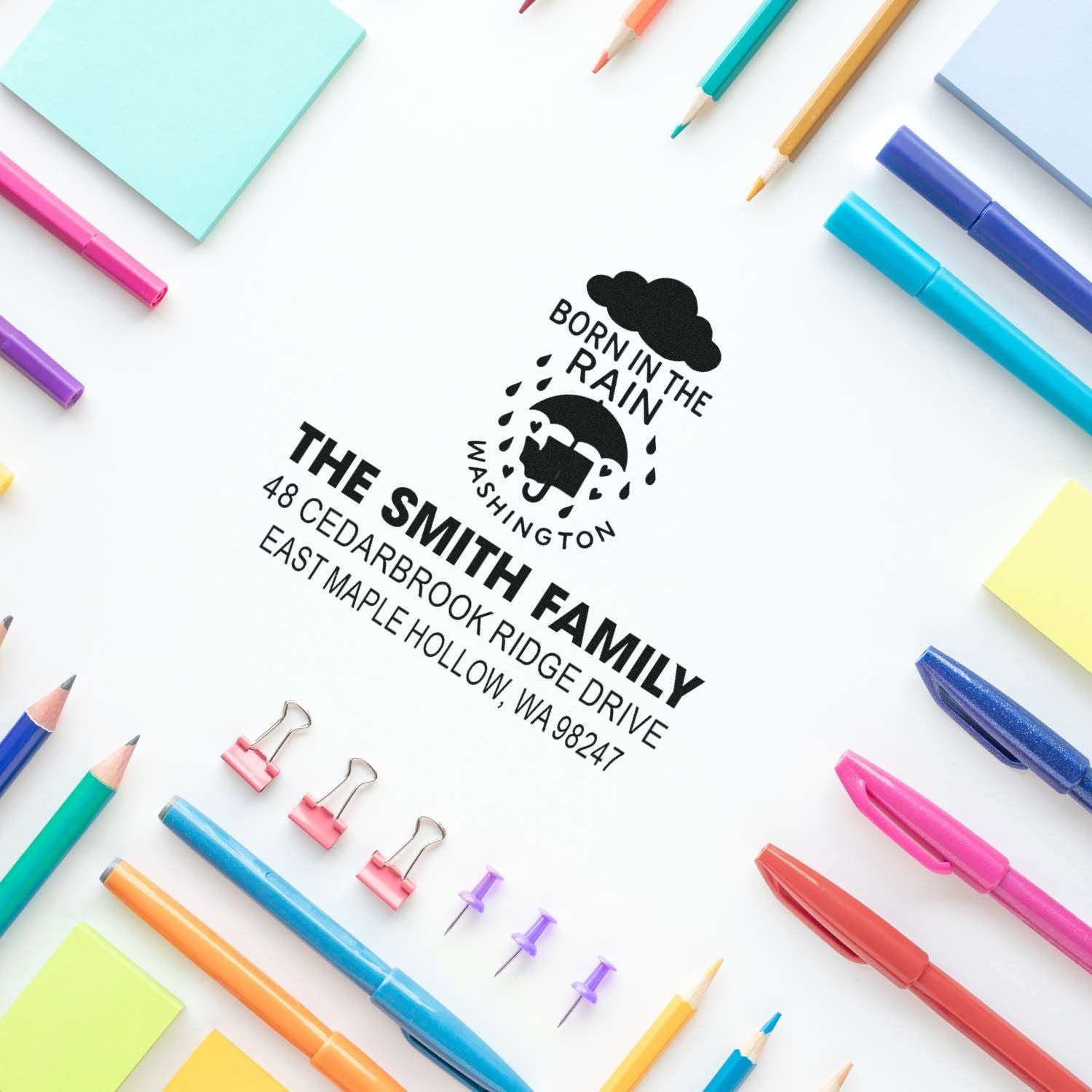 Custom self-inking family address stamp with rain cloud design on white desk, surrounded by colorful pens, pencils, sticky notes, and binder clips.