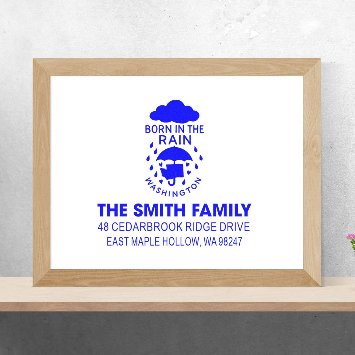 Personalized blue family address stamp for The Smith Family, framed on a shelf against a neutral wall, featuring Born in the Rain Washington and mailing address.