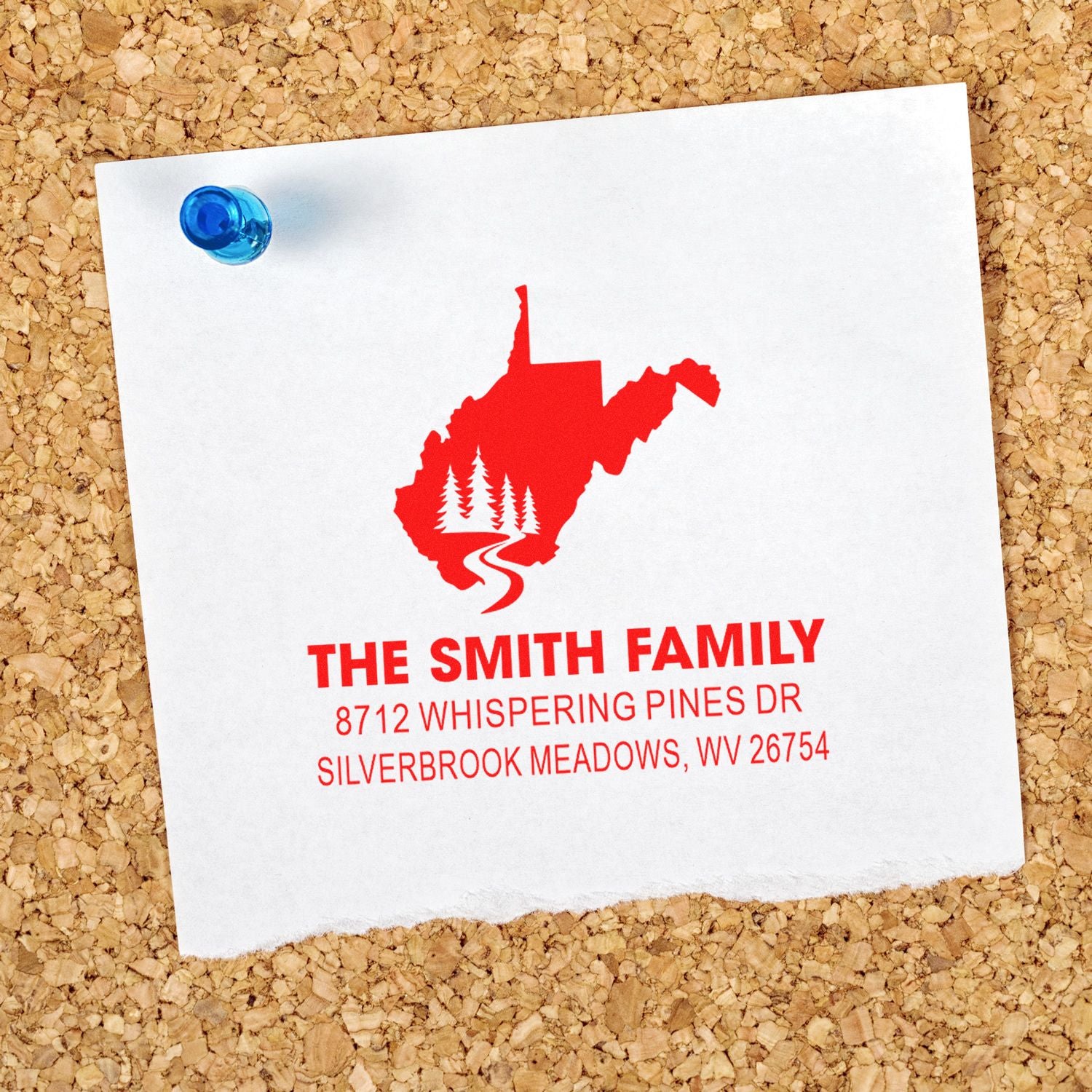 Custom self-inking family address stamp on white paper pinned to corkboard, featuring a West Virginia silhouette with pine trees and road.