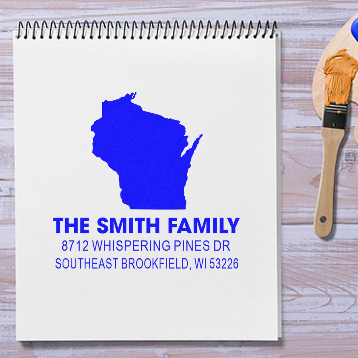 Personalized family address stamp with Wisconsin silhouette on spiral notebook, beside paintbrush on rustic wood desk.