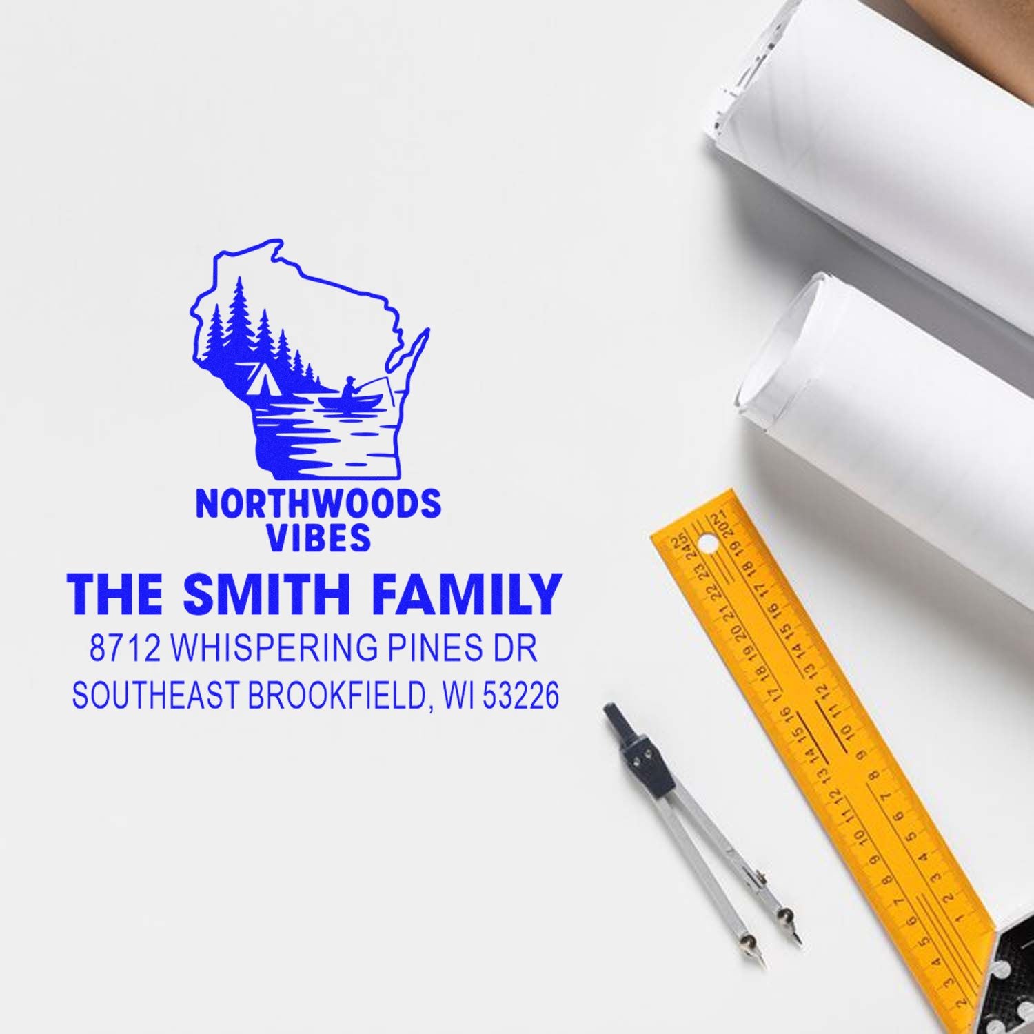 Custom blue Northwoods Vibes family address stamp with Wisconsin outline, on a white desk with ruler, compass, and packaging.