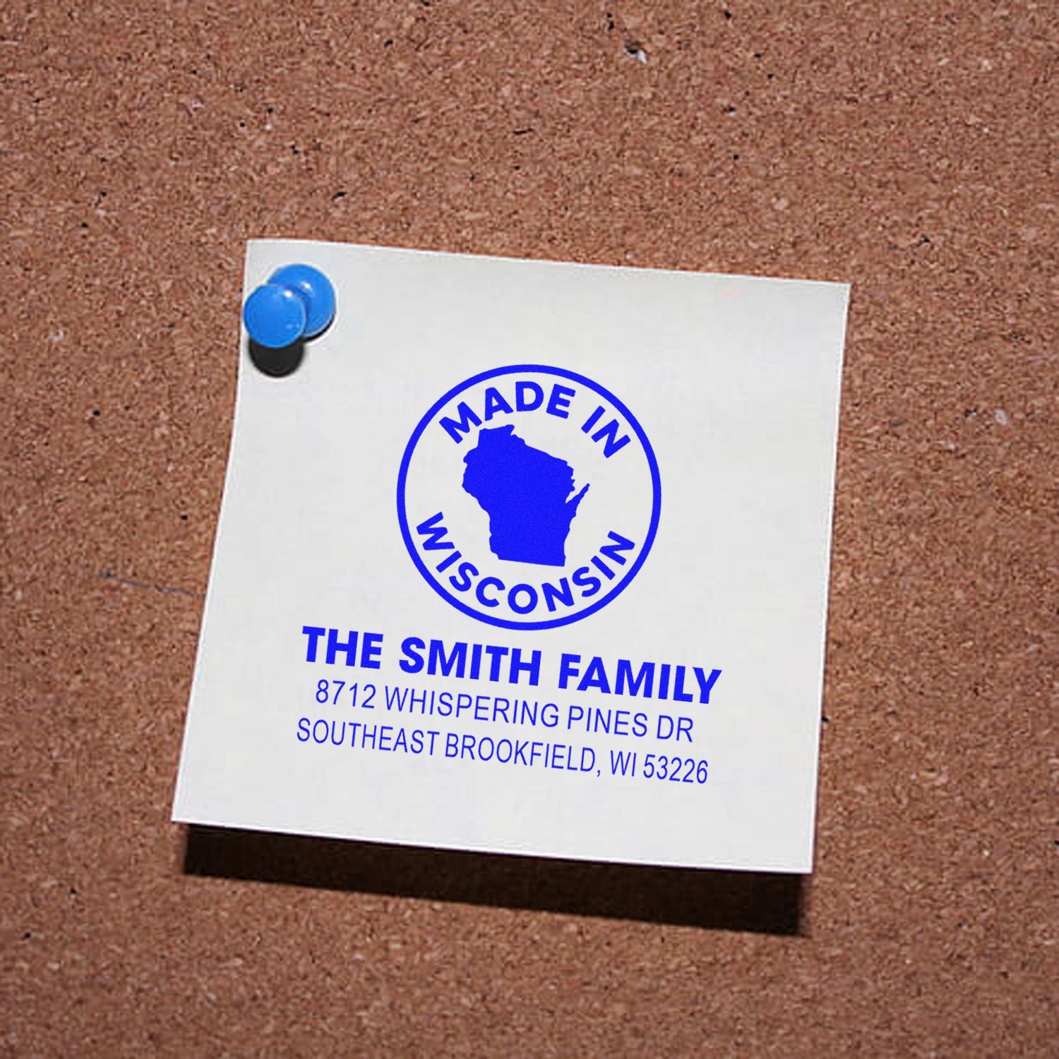 Custom Made in Wisconsin address stamp for The Smith Family, blue imprint on white note pinned to a corkboard with a blue pushpin.
