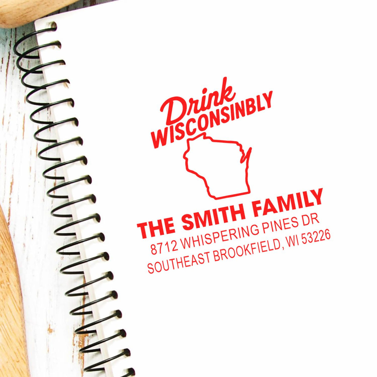 Custom red self-inking address stamp on a spiral notebook, featuring Drink Wisconsinbly, Wisconsin outline, and the Smith family address.