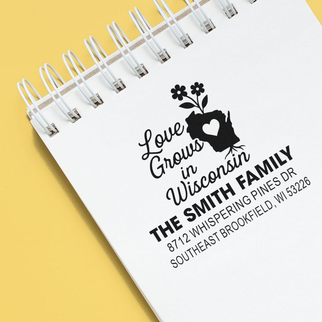 Custom return address stamps on a spiral notebook, featuring a floral Wisconsin family logo and bold name on a yellow background.