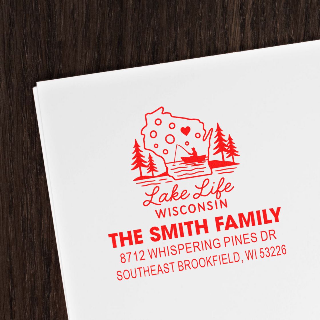 Custom red return address stamp with Wisconsin lake life design, family name and address on white paper beside a dark wood surface.