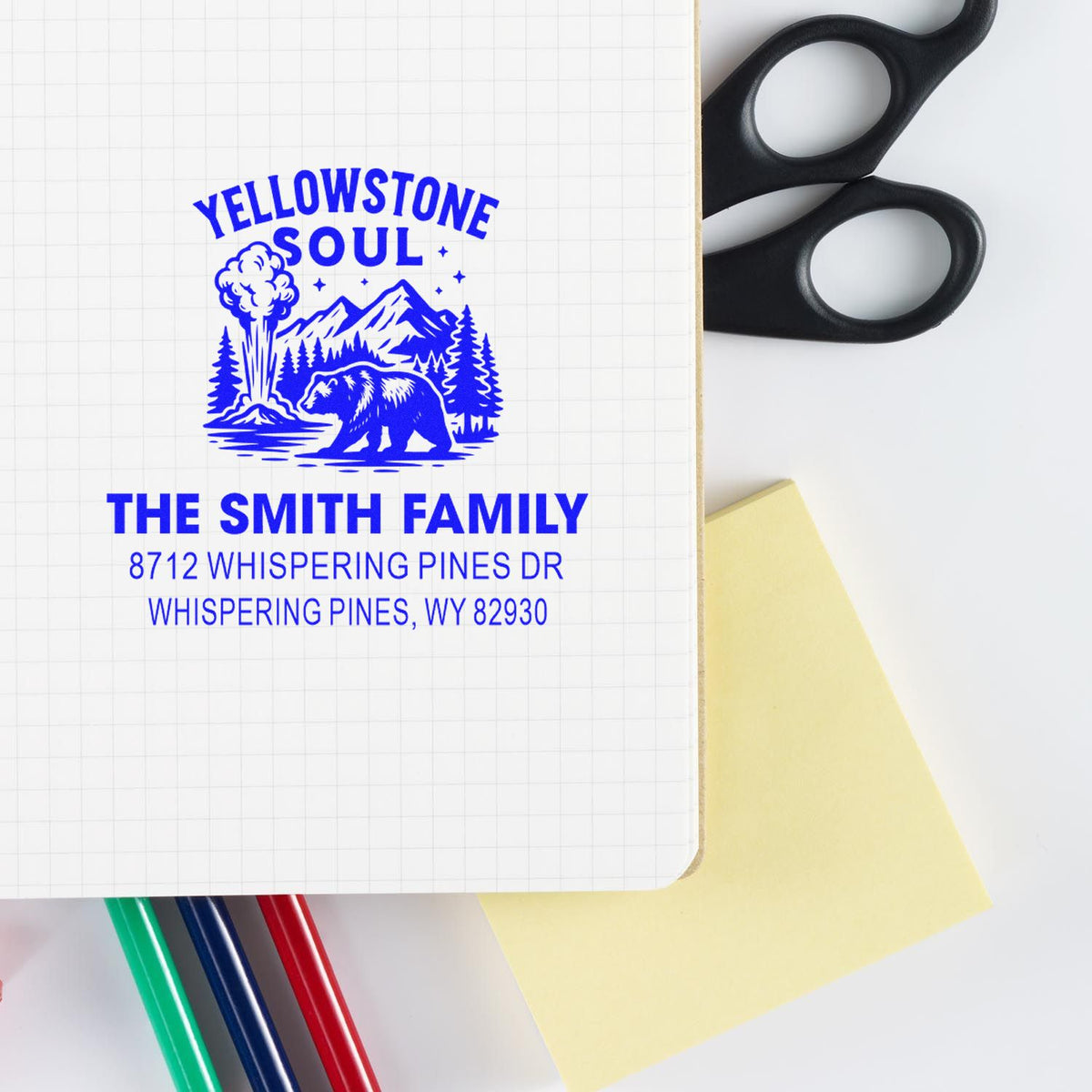 Custom Yellowstone Soul family address stamp impression on notebook paper, with bear, mountains and geyser design beside scissors, sticky note and pens.