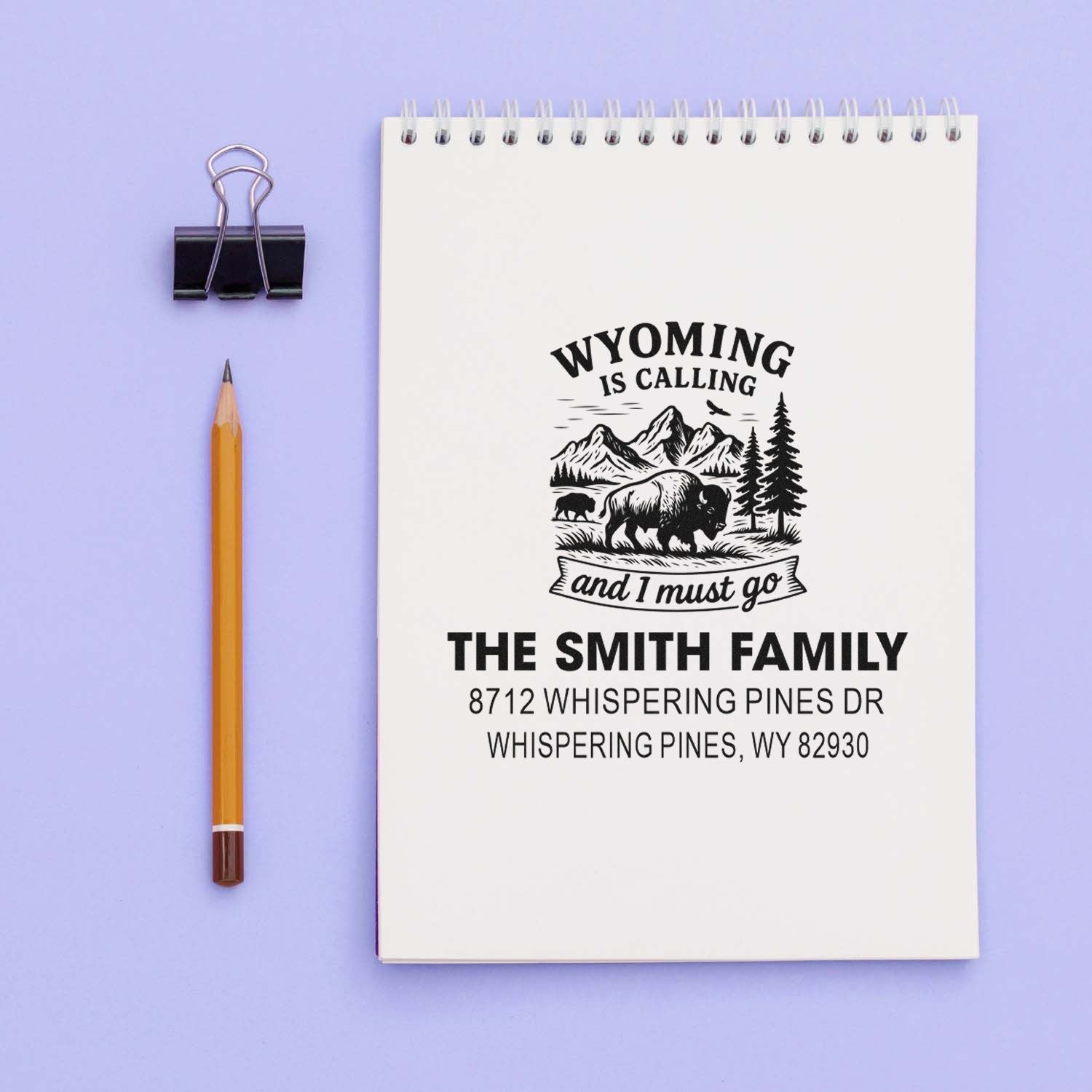 Custom Wyoming family address stamp on a spiral notebook, with bison and mountain design, beside a pencil and binder clip on lavender background.