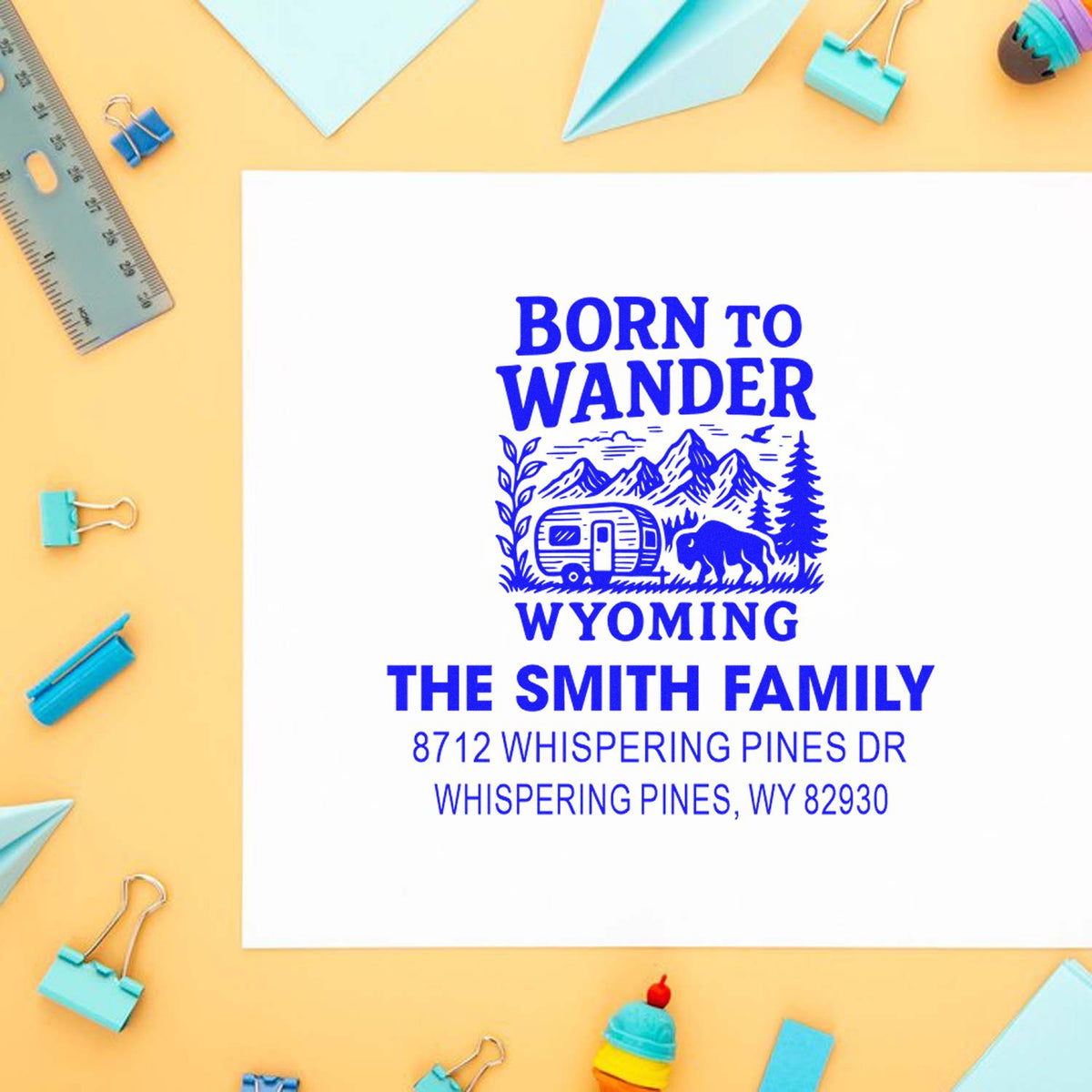 Custom Wyoming return address stamp with Born to Wander, mountains, camper and bison, displayed on white paper with pastel stationery on a desk.