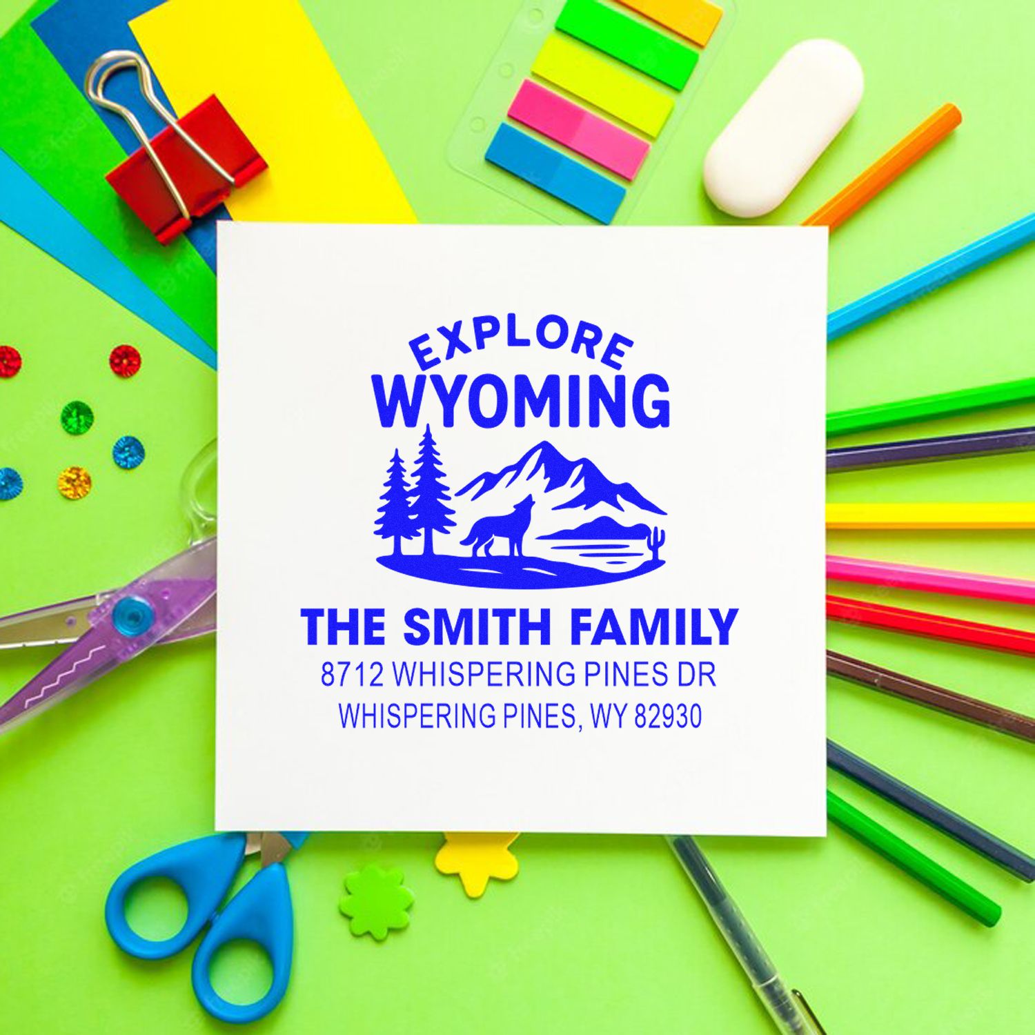 Personalized Wyoming address stamp with mountain and pine design, surrounded by colorful stationery, paper clips, scissors, pens, and craft supplies.