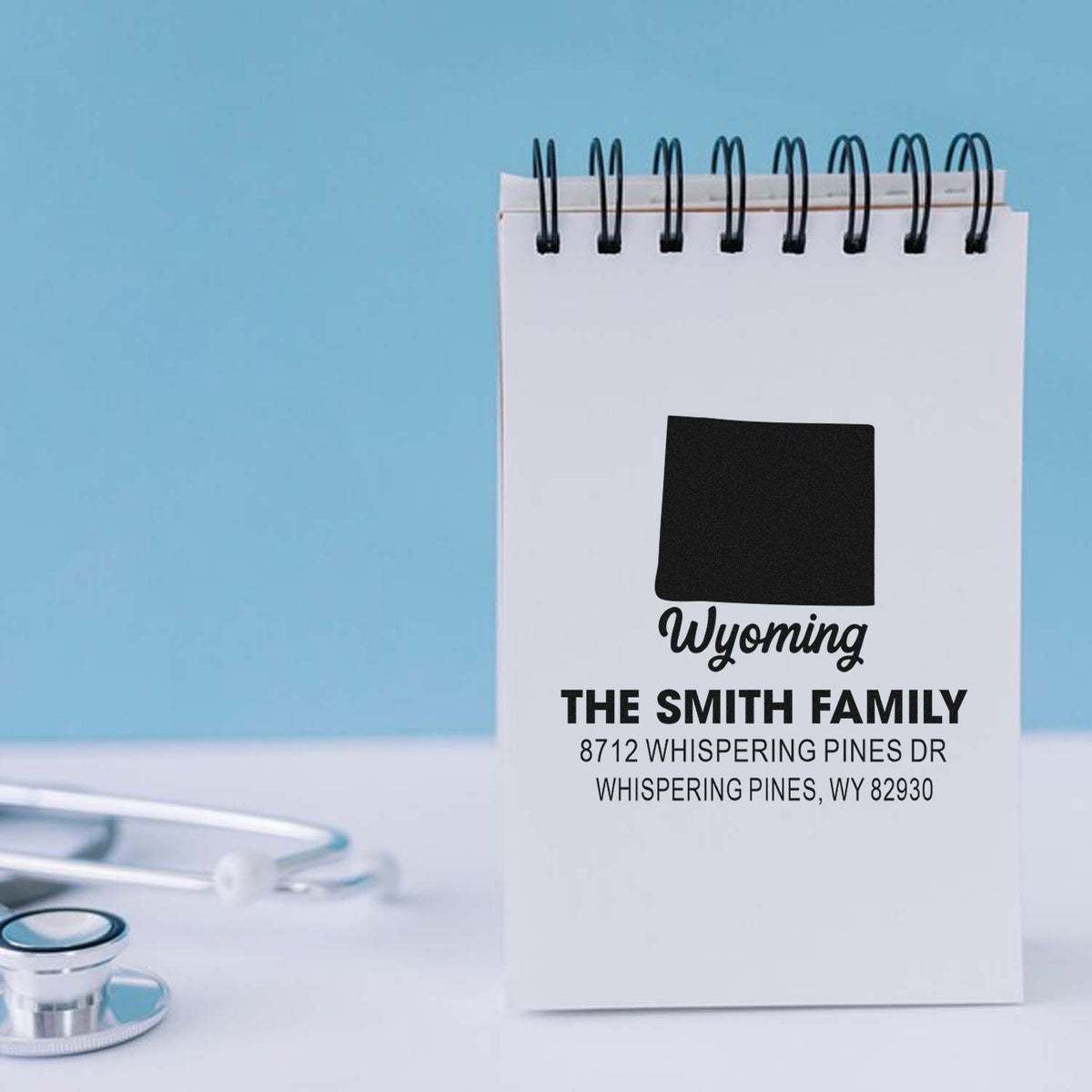 Custom Wyoming address stamp on a spiral notepad, showing family name and mailing address, with a stethoscope on a clean blue desk.