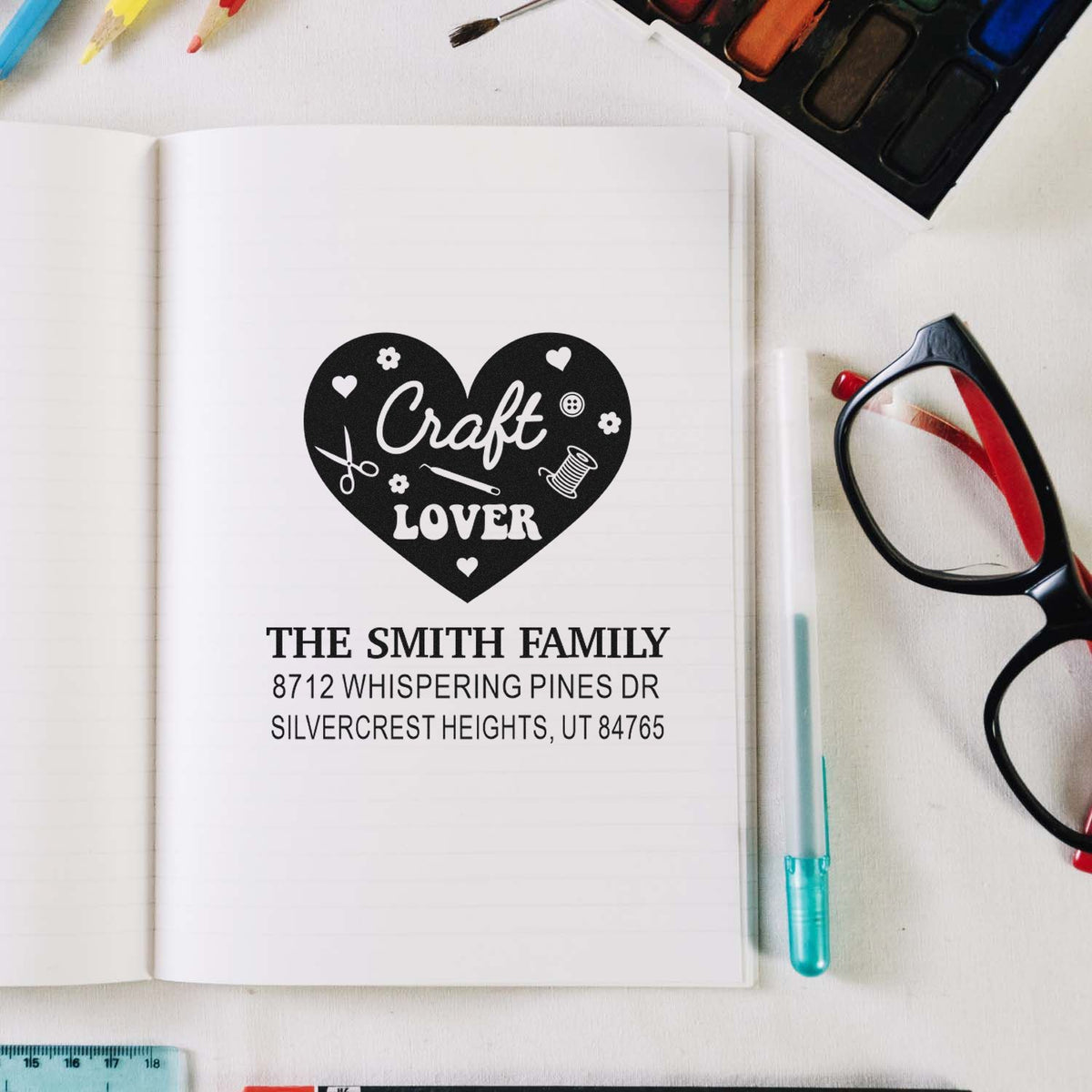 Personalized craft lover address stamp on an open notebook, featuring a heart design and family address, surrounded by stationery, glasses and paints.