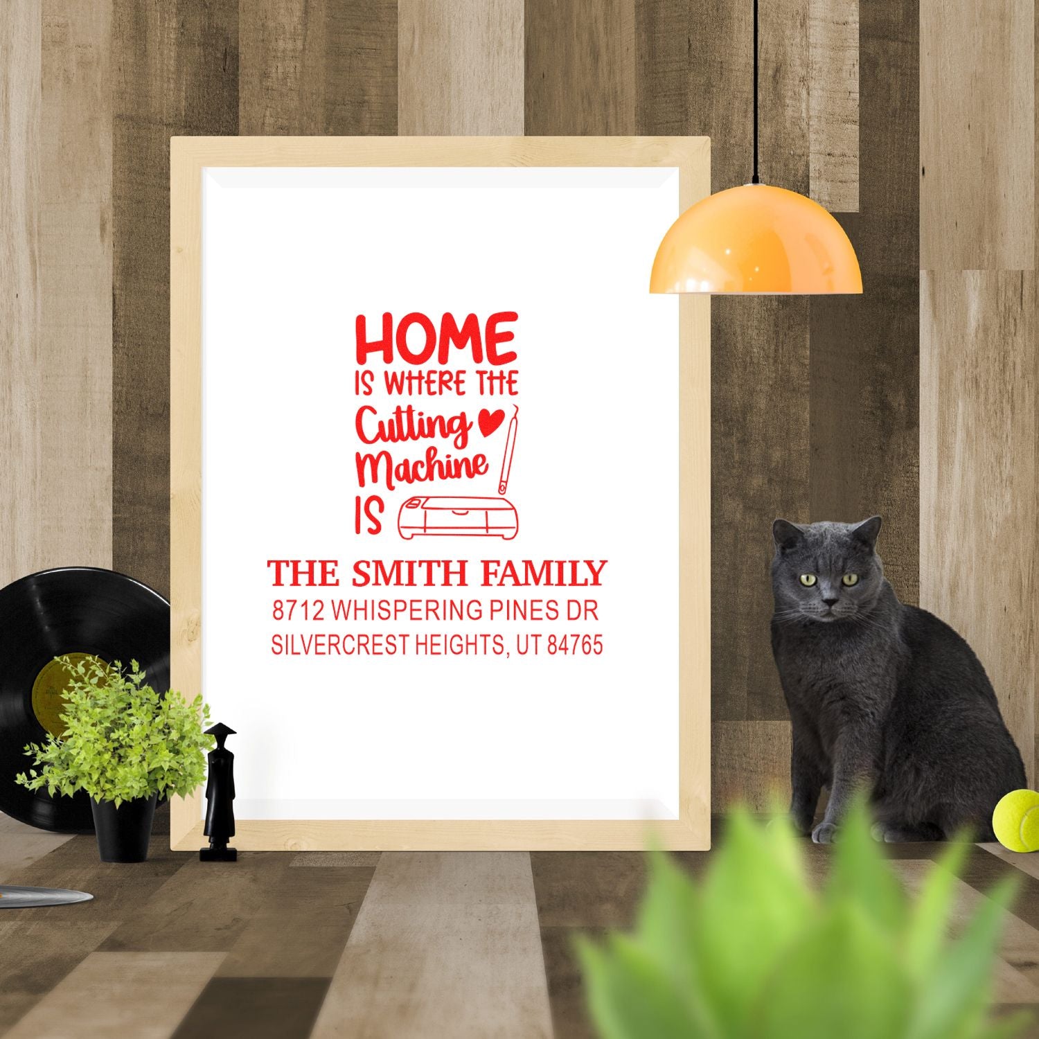 Personalized home stamp design with crafting machine, family name and address in a framed display, styled with a cat, lamp, plants and tennis ball.