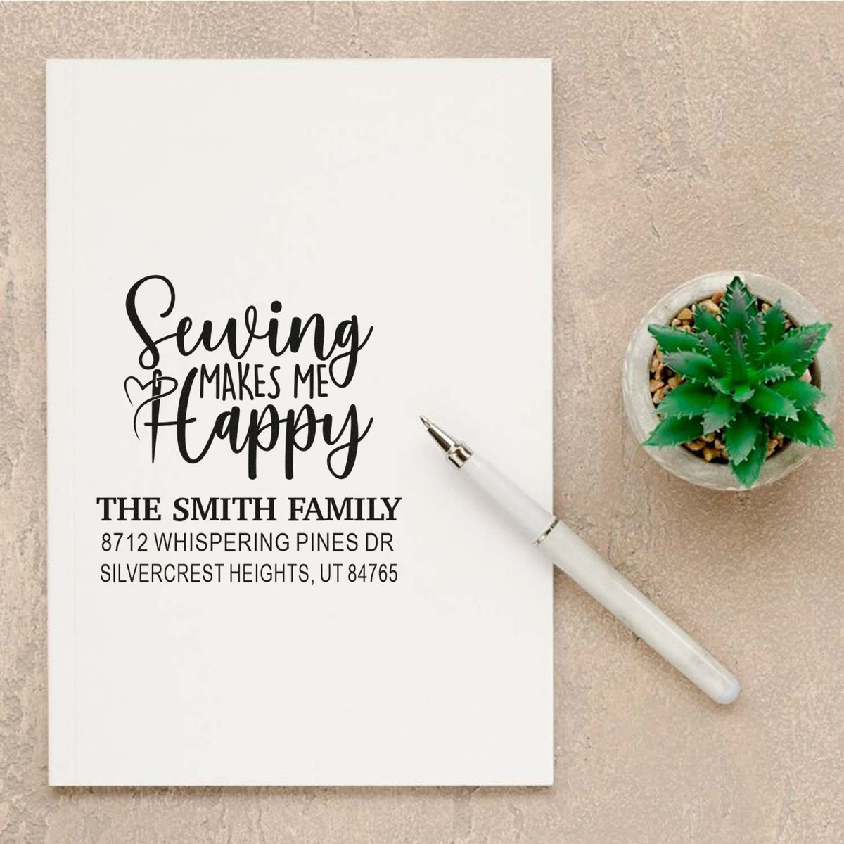 Custom return address stamp with Seuvin Makes Me Happy on paper, beside a white pen and potted succulent.