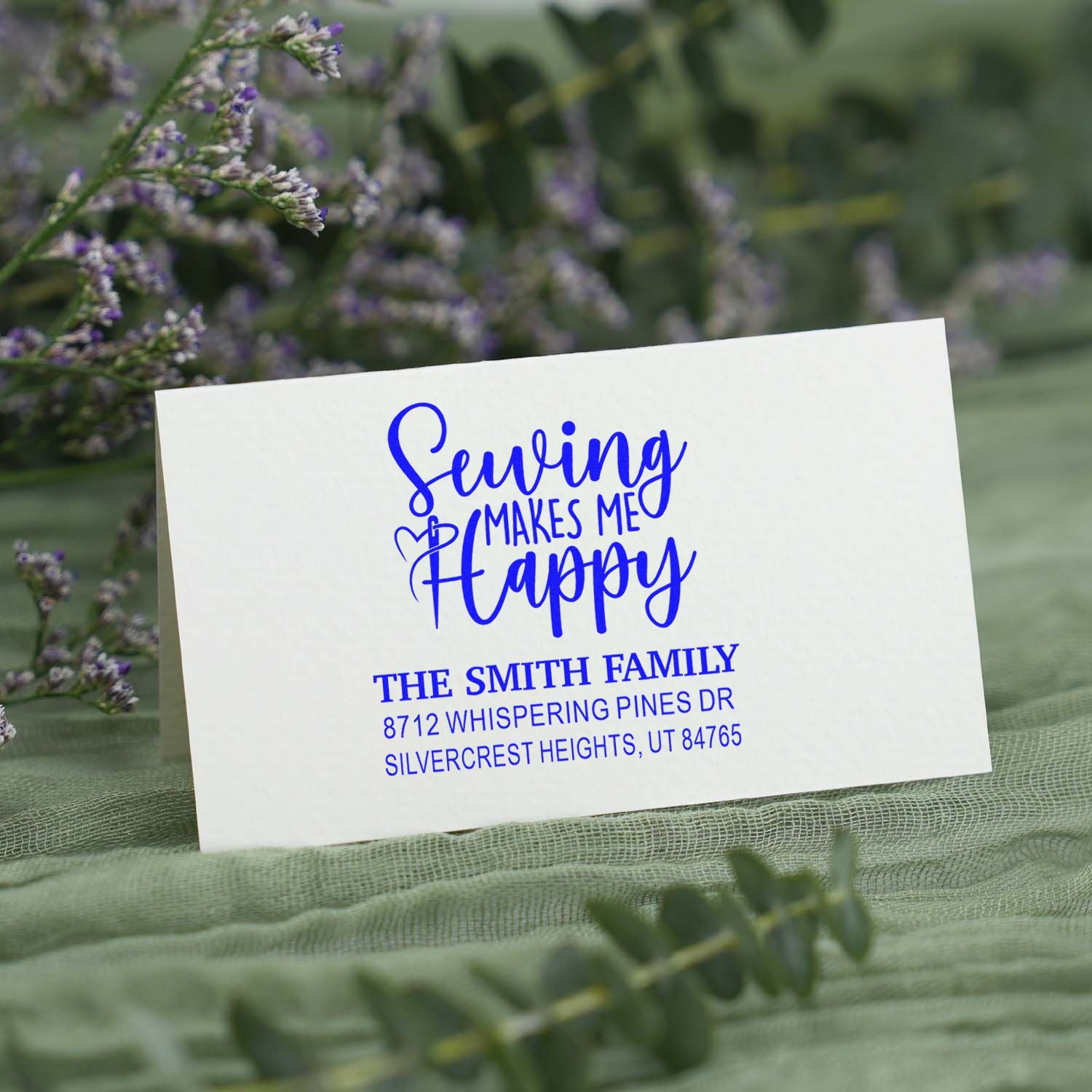 Custom blue self-inking address stamp with Sewing Makes Me Happy on white card, styled on green fabric with purple flowers.