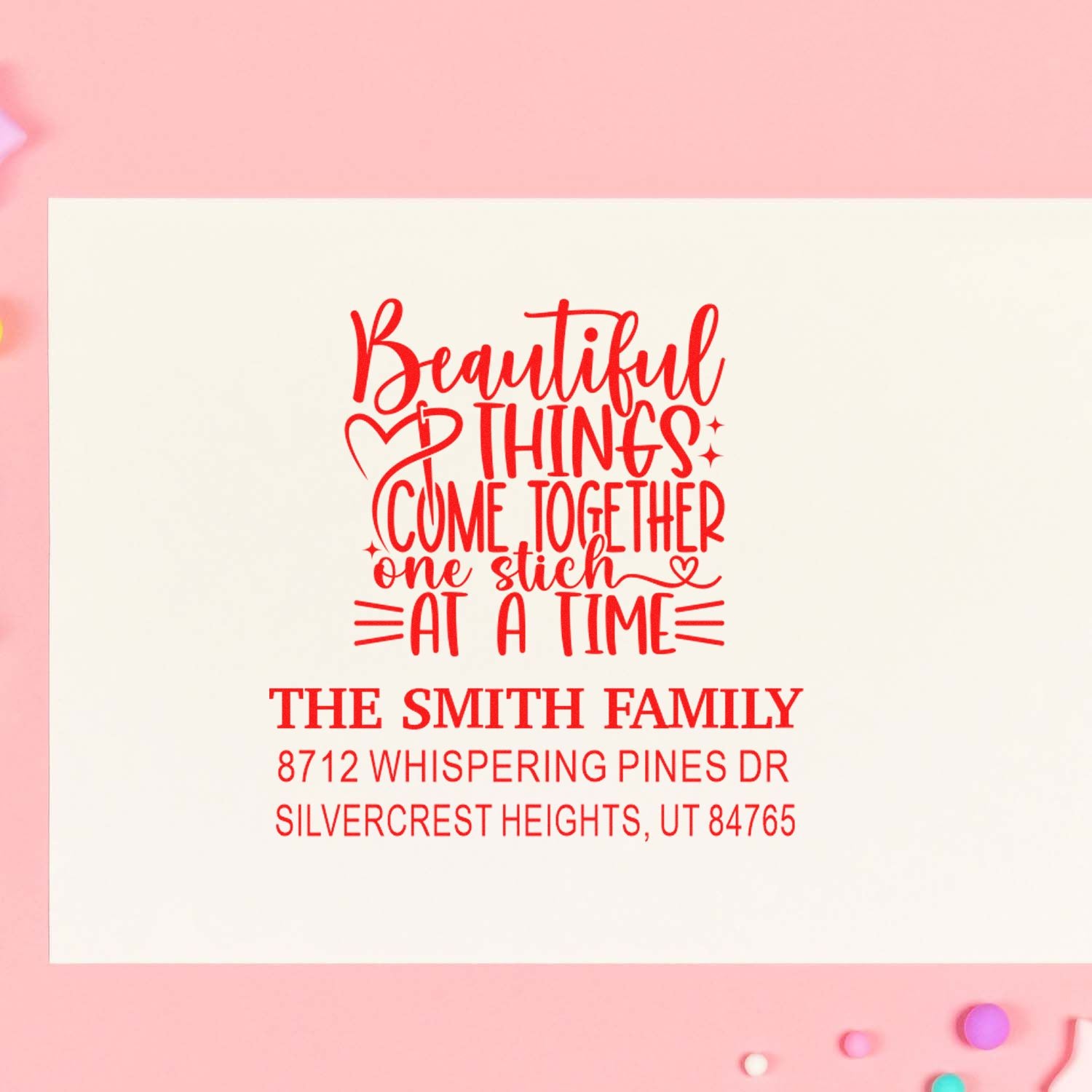 Personalized family address stamp with Beautiful Things Come Together One Stitch at a Time quote on cream card against pink background.