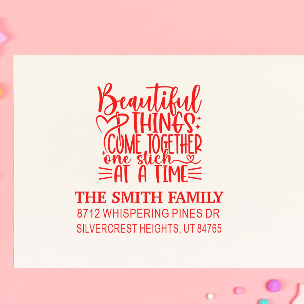Red custom return address stamp with Beautiful things come together one stitch at a time for The Smith Family on a white card, pink background.