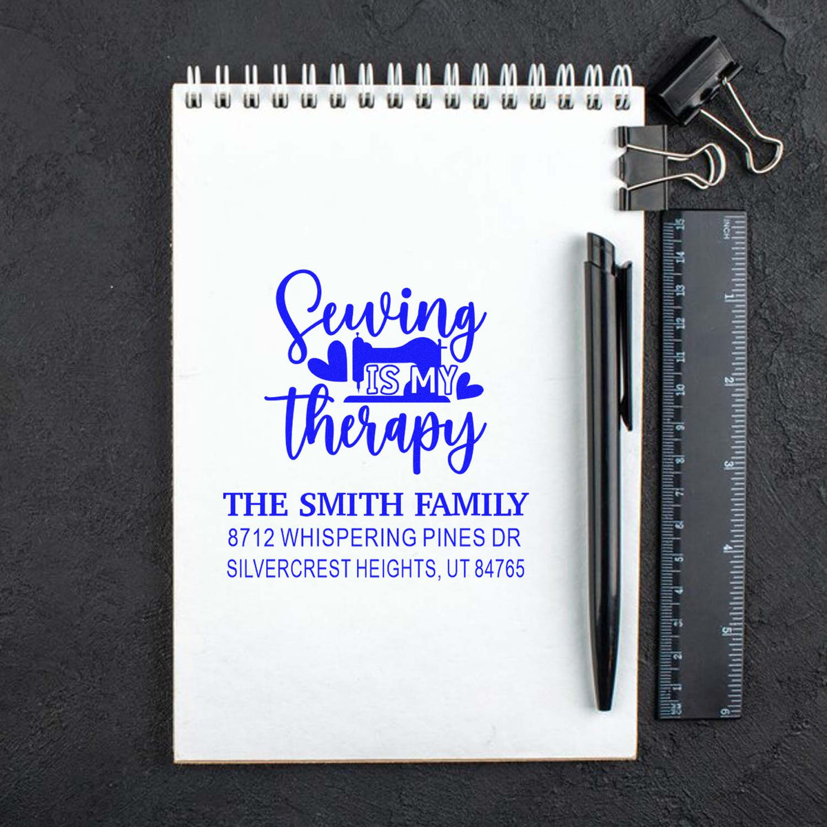 Custom blue self-inking stamp on spiral notebook reading Sewing is my therapy, with family name and address, beside pen, ruler, and binder clips.