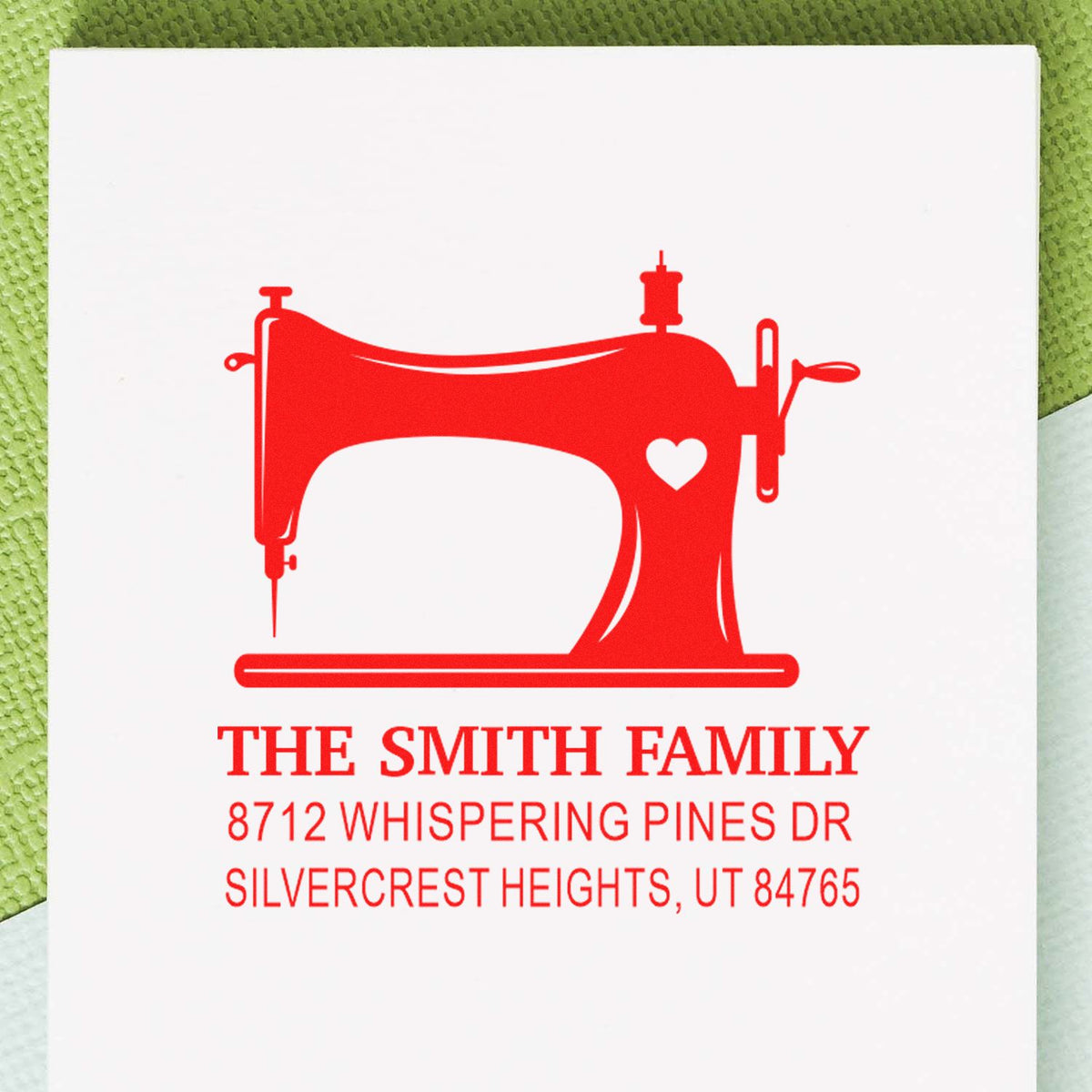 Red personalized address stamp with sewing machine and heart design for The Smith Family, on white card against a green textured background.