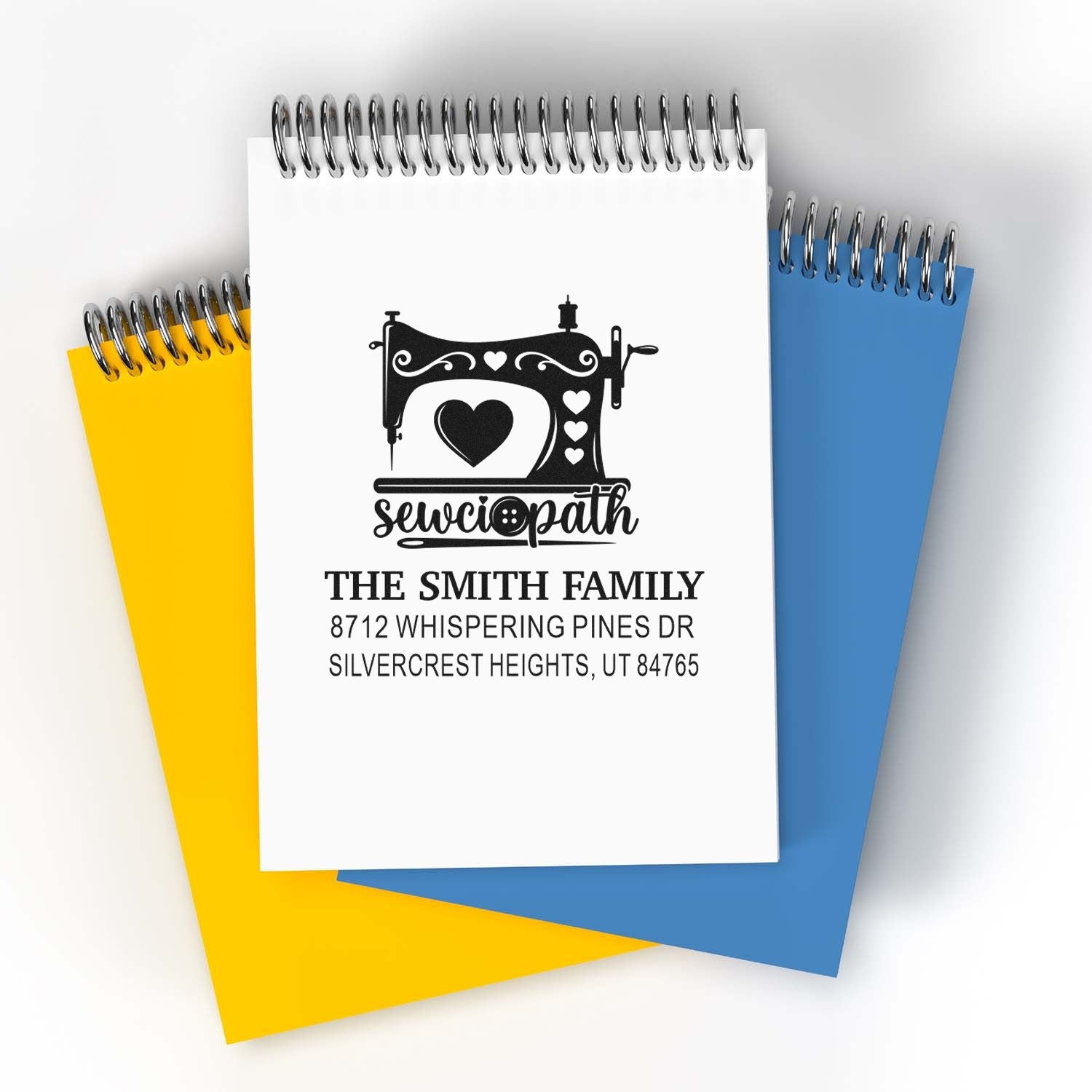 Custom address stamp featuring a sewing machine and heart on a spiral notebook, layered over yellow and blue notebooks with family name and address.