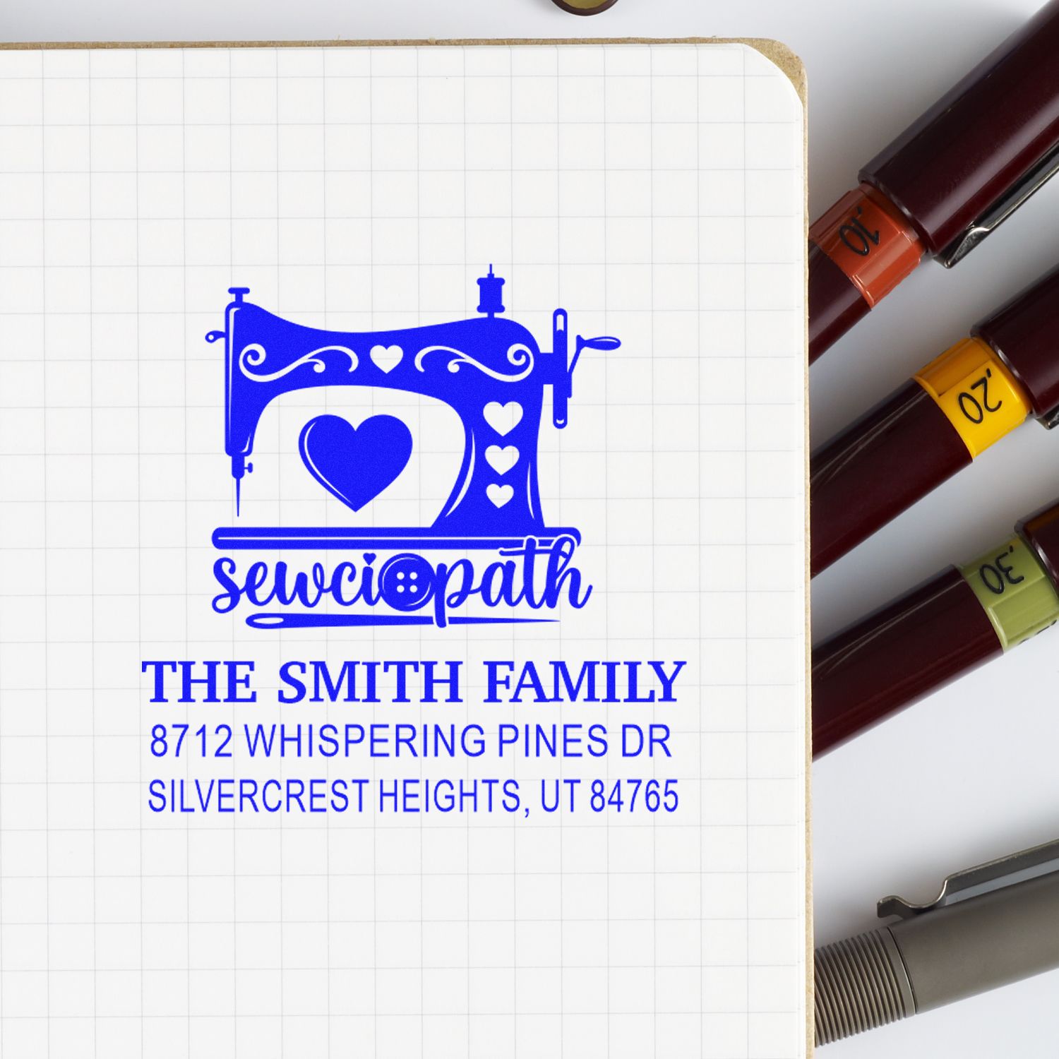 Custom address stamp with sewing machine and heart design for The Smith Family, stamped in blue on notebook beside ink stamp markers.