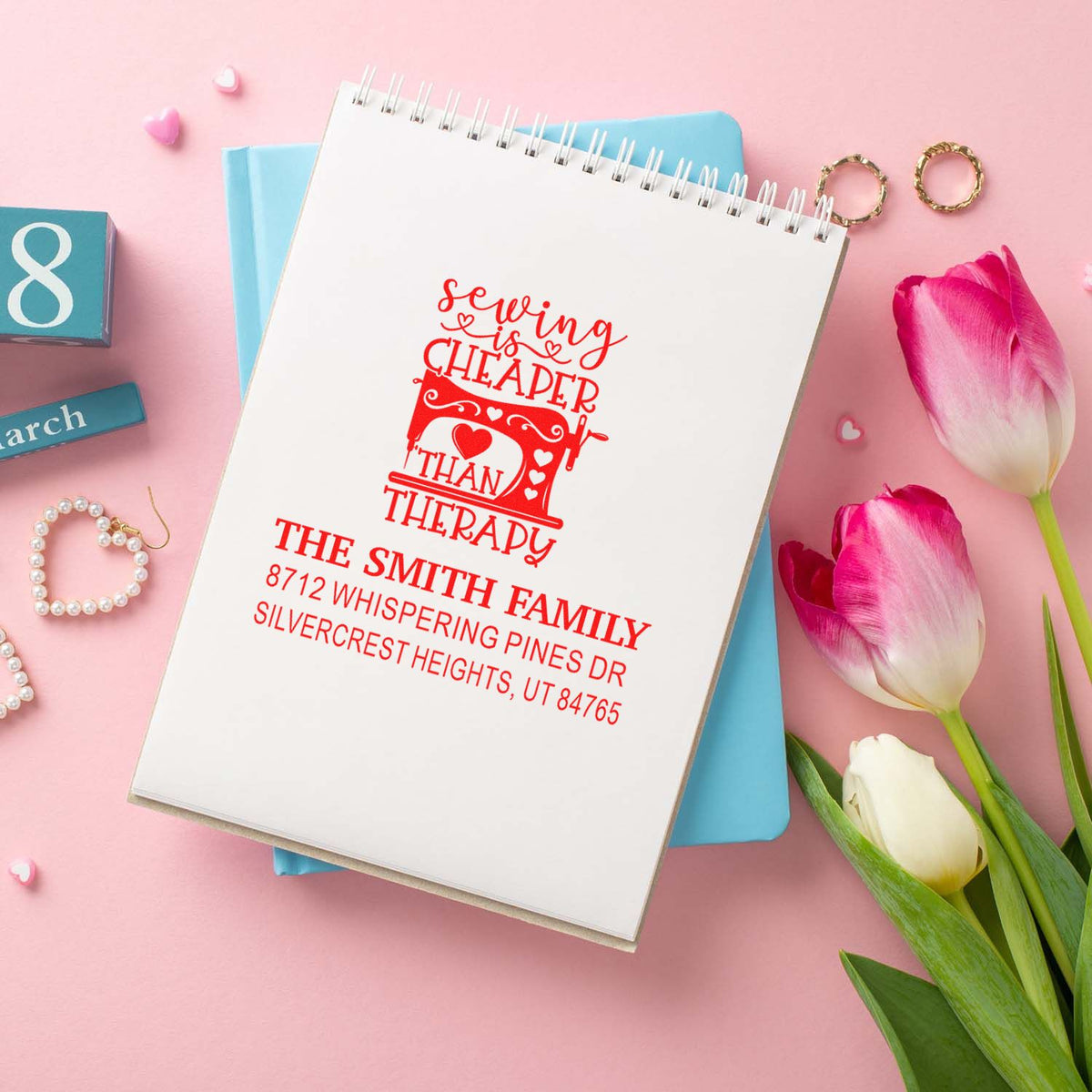 Personalized red sewing-themed address stamp on a spiral notebook, styled with pink tulips, rings, and pastel decor on a pink background.
