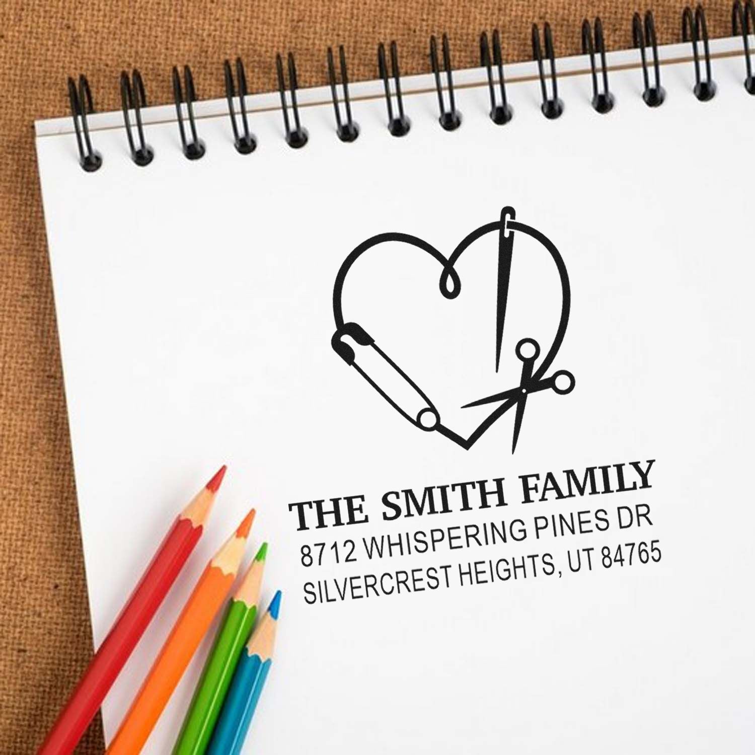 Custom family address stamp with heart sewing motif on a spiral notebook, shown with colored pencils on a desk.