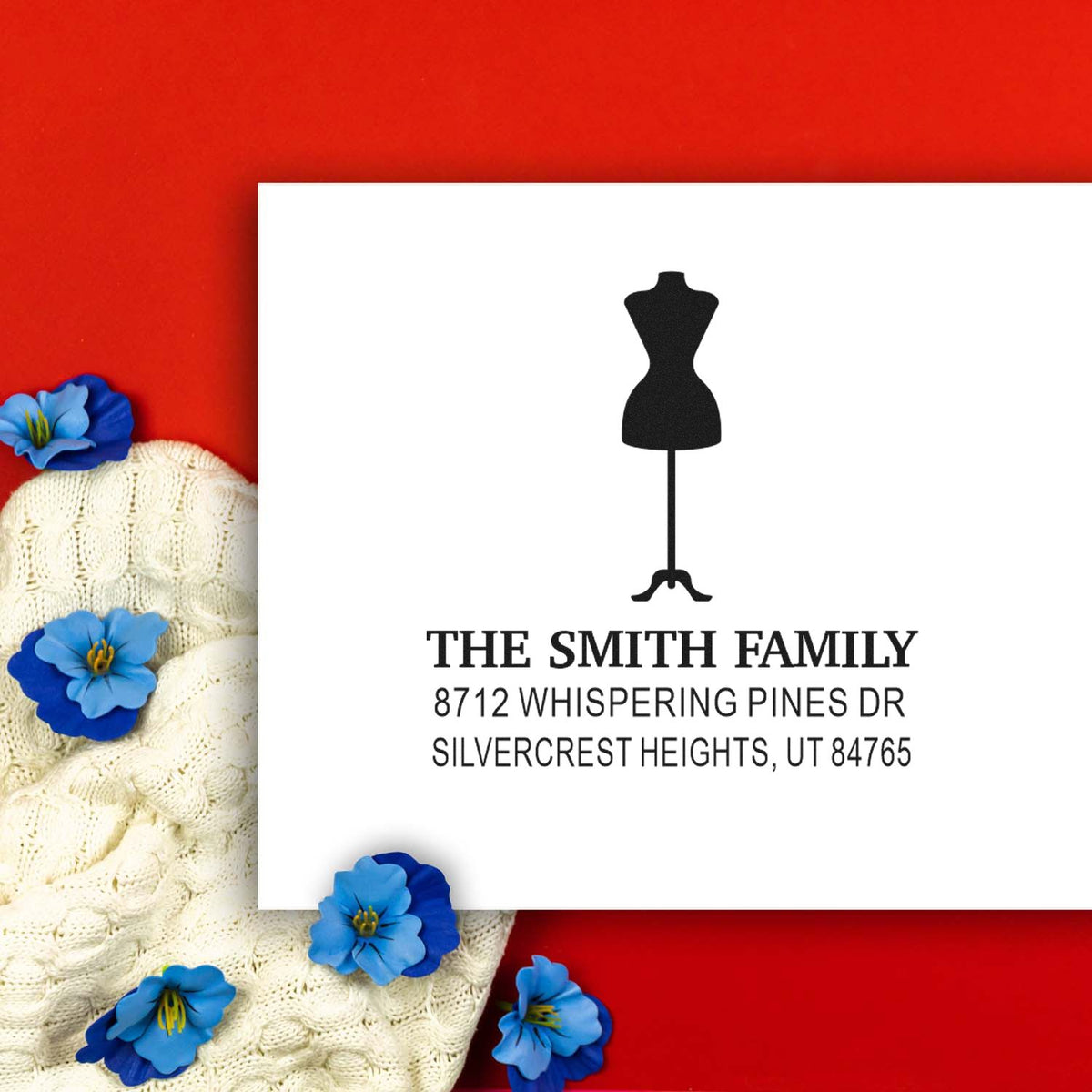 Custom return address stamp with dress form design and The Smith Family address on a white card, styled on a red background with knit sweater and blue flowers.