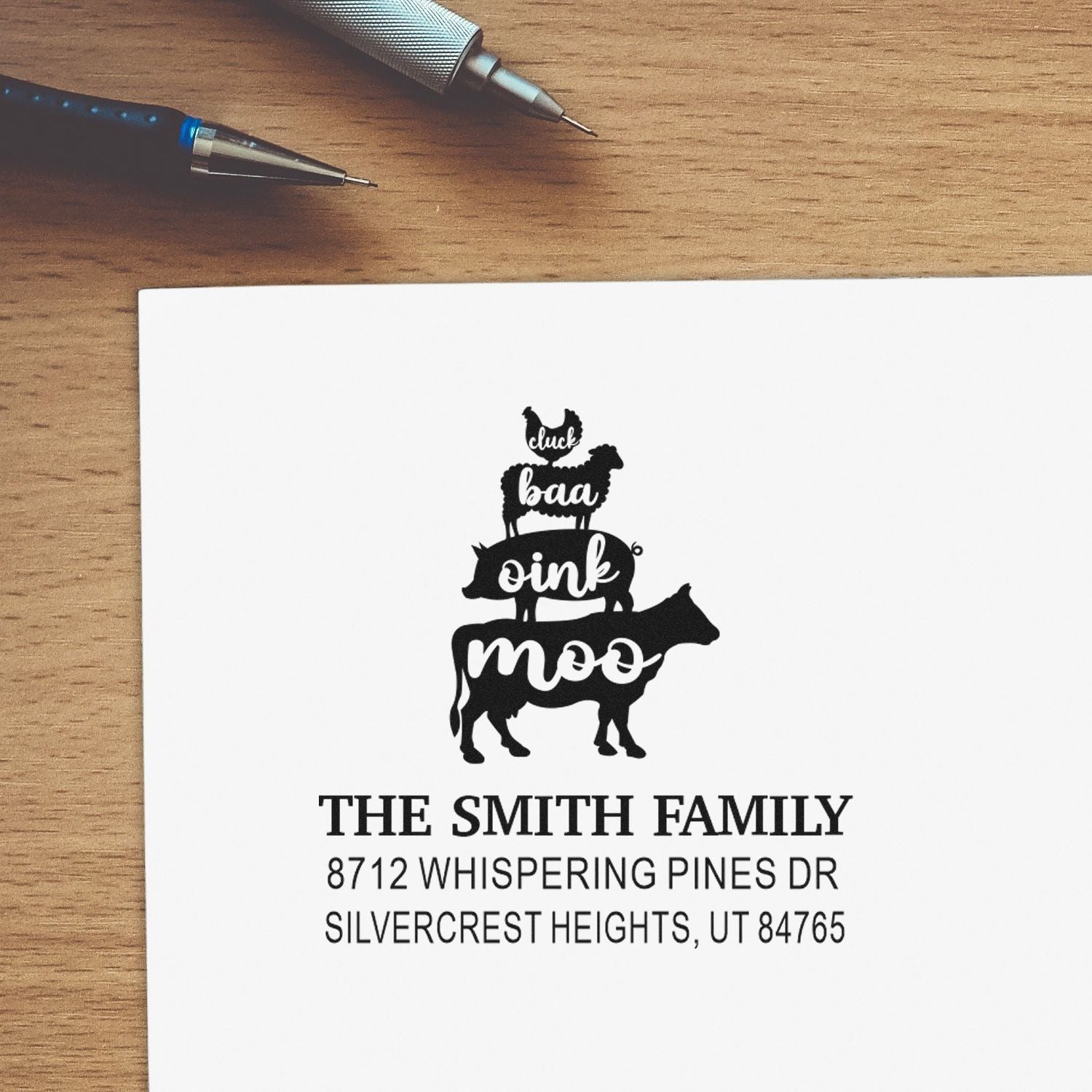 Custom family return address stamp with stacked farm animals saying cluck, baa, oink, moo on white paper beside pens on a wooden desk.
