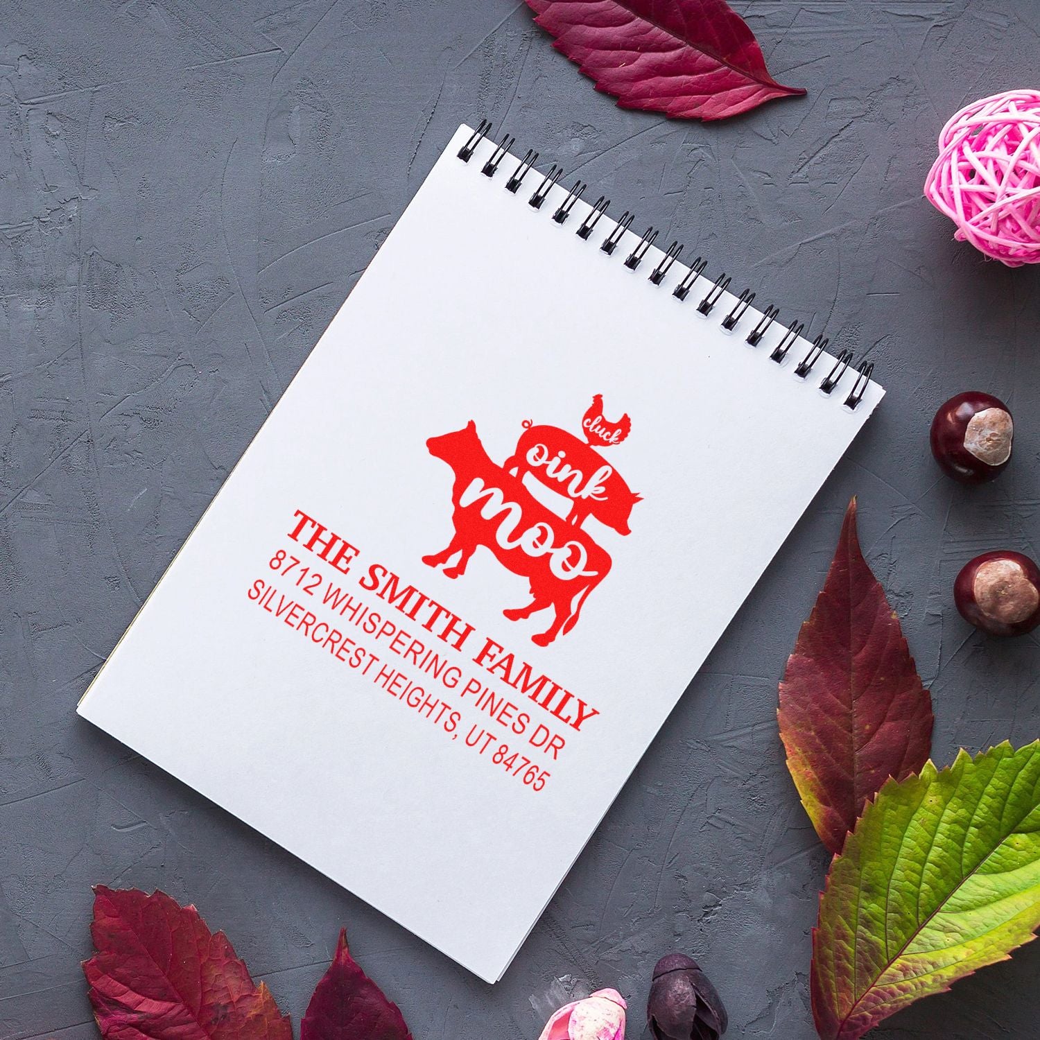 Custom red return address stamp on spiral notebook, with pink cow design, set on gray tabletop with autumn leaves and decorative accents.