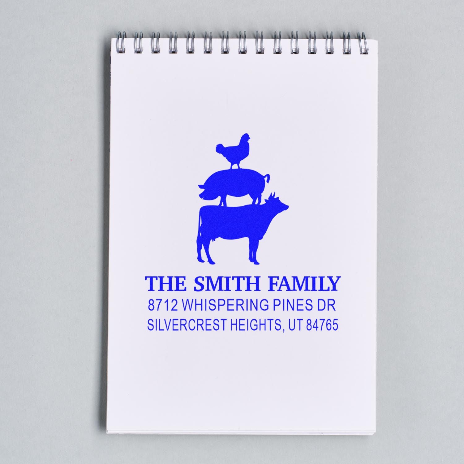 Custom blue address stamp with stacked chicken, pig and cow design on a spiral notebook, reading THE SMITH FAMILY and Silvercrest Heights, UT.