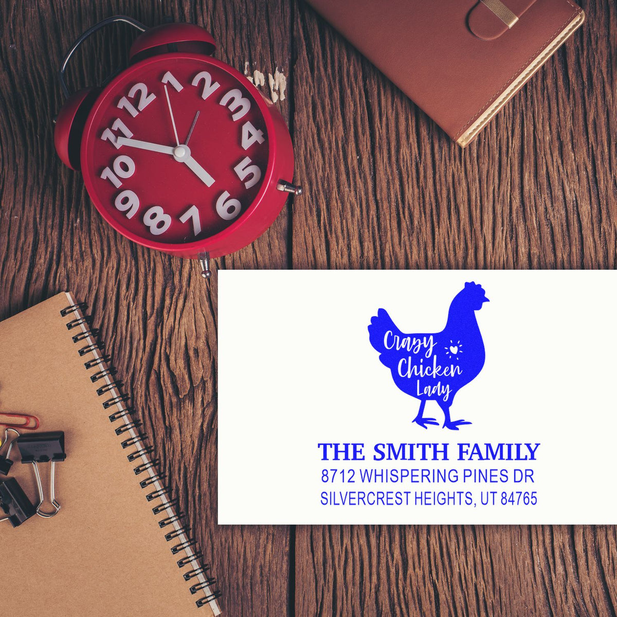 Custom self-inking address stamp with blue chicken design and The Smith Family address, displayed on a white card on a rustic wood desk with notebook and clock.