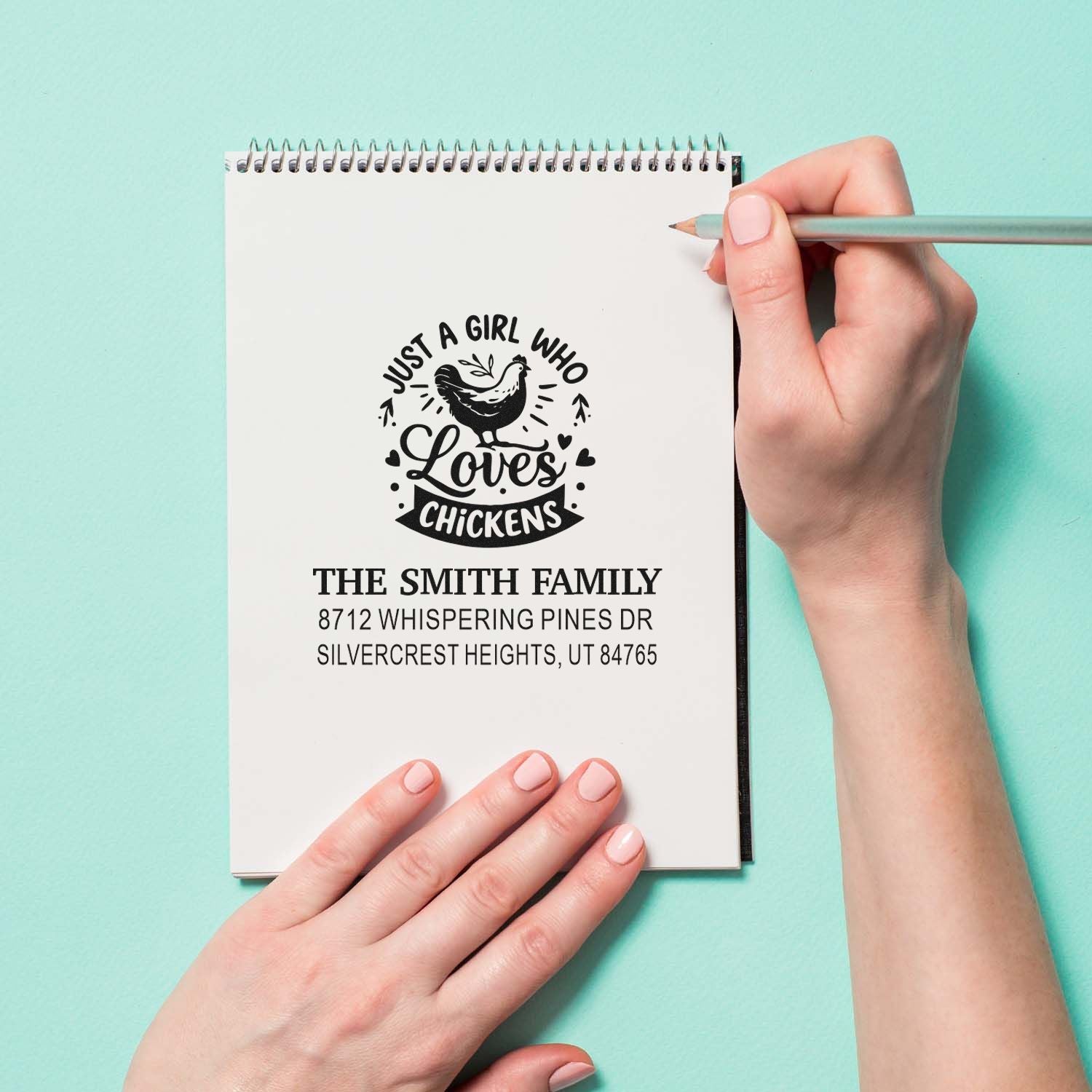 Hand using a pencil beside a spiral notebook featuring a custom chicken-loving family return address stamp on a mint background.