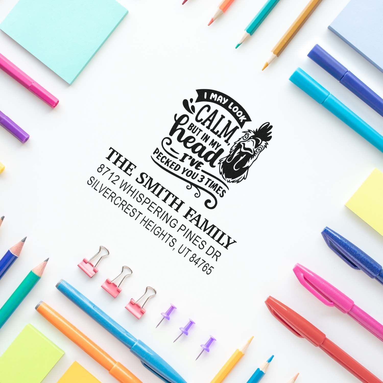 Custom address stamp impressions on white paper with a rooster quote and The Smith Family return address, framed by colorful pens, pencils, and notes.