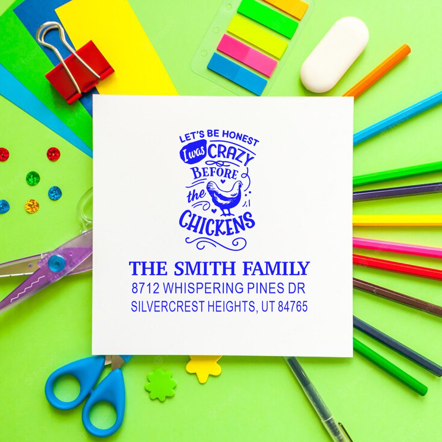 Personalized address stamp with funny chicken design on white card, surrounded by colorful office supplies, scissors, clips, and markers.