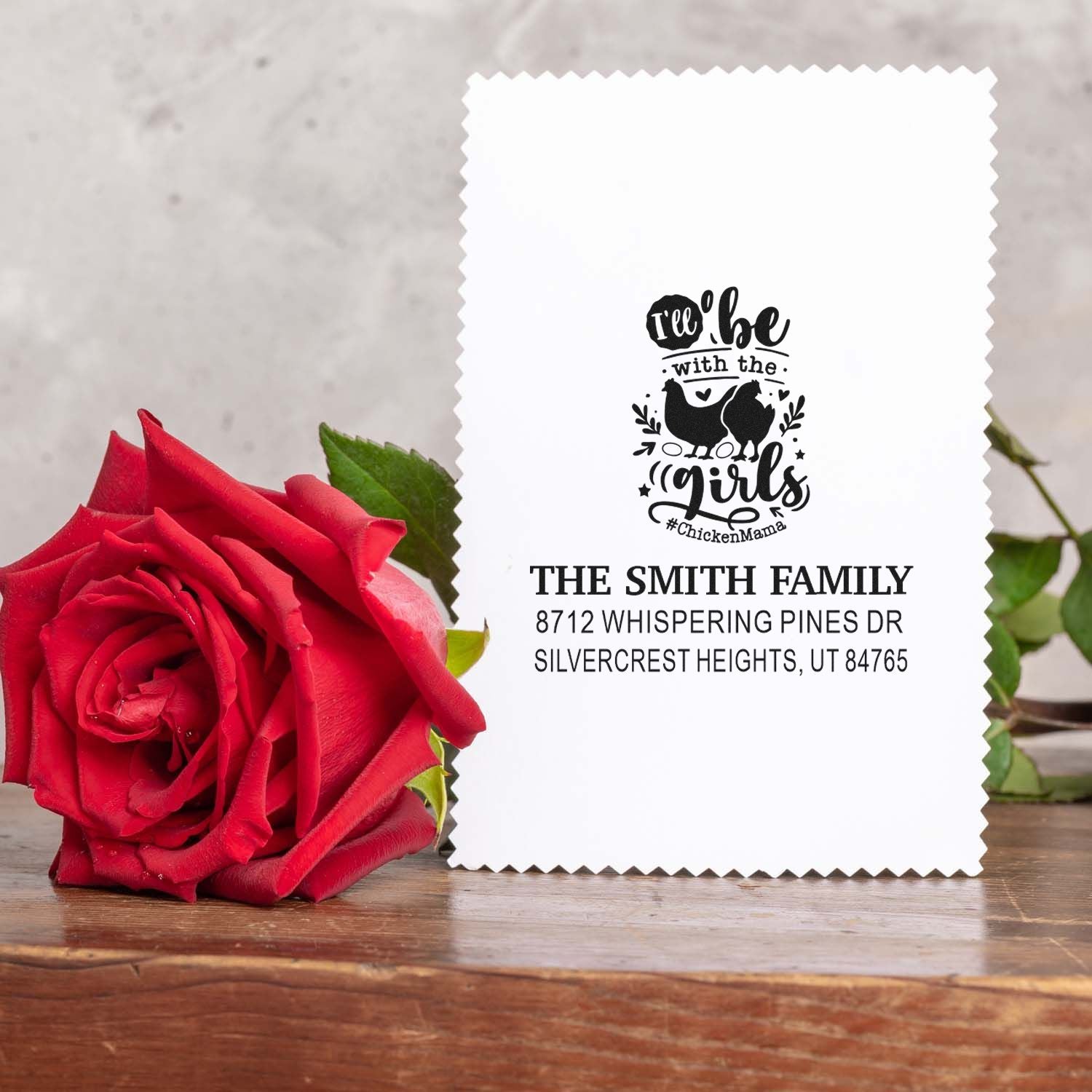 Personalized address stamp with Ill be with the girls chicken design, displayed beside a red rose on a rustic wooden table.