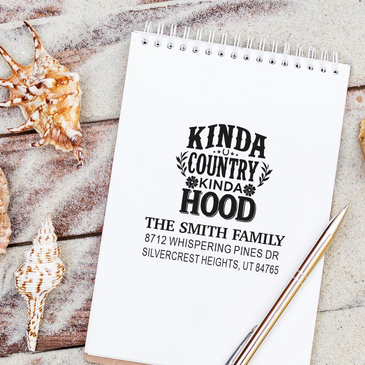 Custom address stamp on spiral notebook reading Kinda Country Kinda Hood, styled with seashells, sand, and a gold pen.