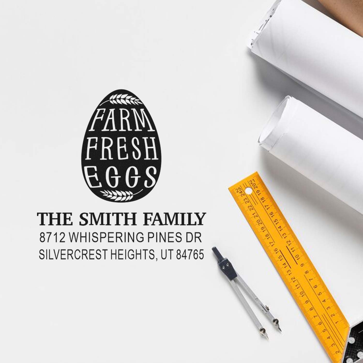 Custom farm fresh eggs address stamp with the Smith Family name and address, shown with rulers and drafting tools on a white tabletop.