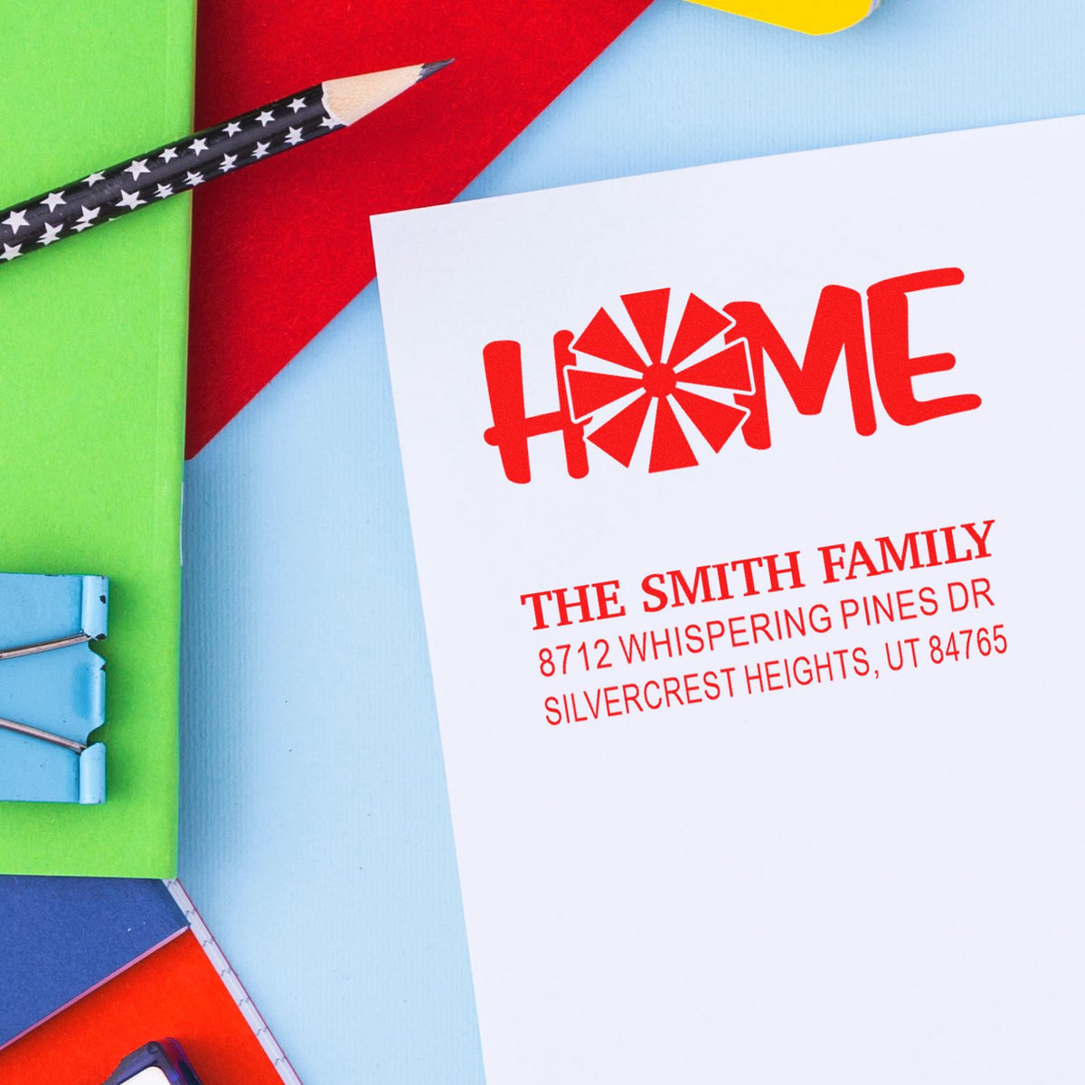 Custom home address stamp with red HOME logo, family name and address, arranged on colorful stationery with pencil and clips.