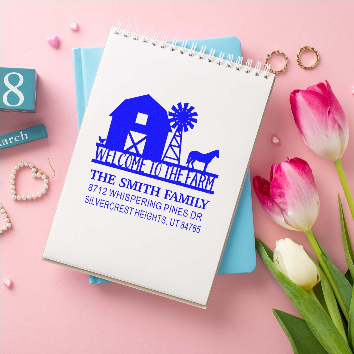 Personalized blue farm welcome stamp on a spiral notebook, featuring a barn, windmill and horse, styled on a pink flat lay with tulips and decor.