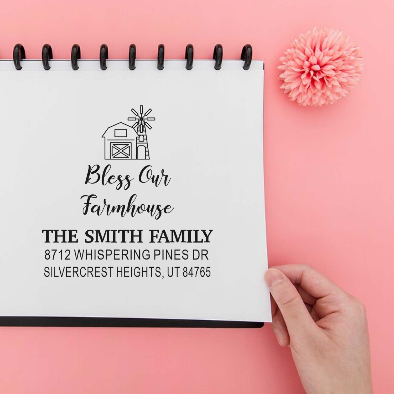 Personalized farmhouse return address stamp in a spiral notebook on a pink background, held by hand with a pink flower accent.