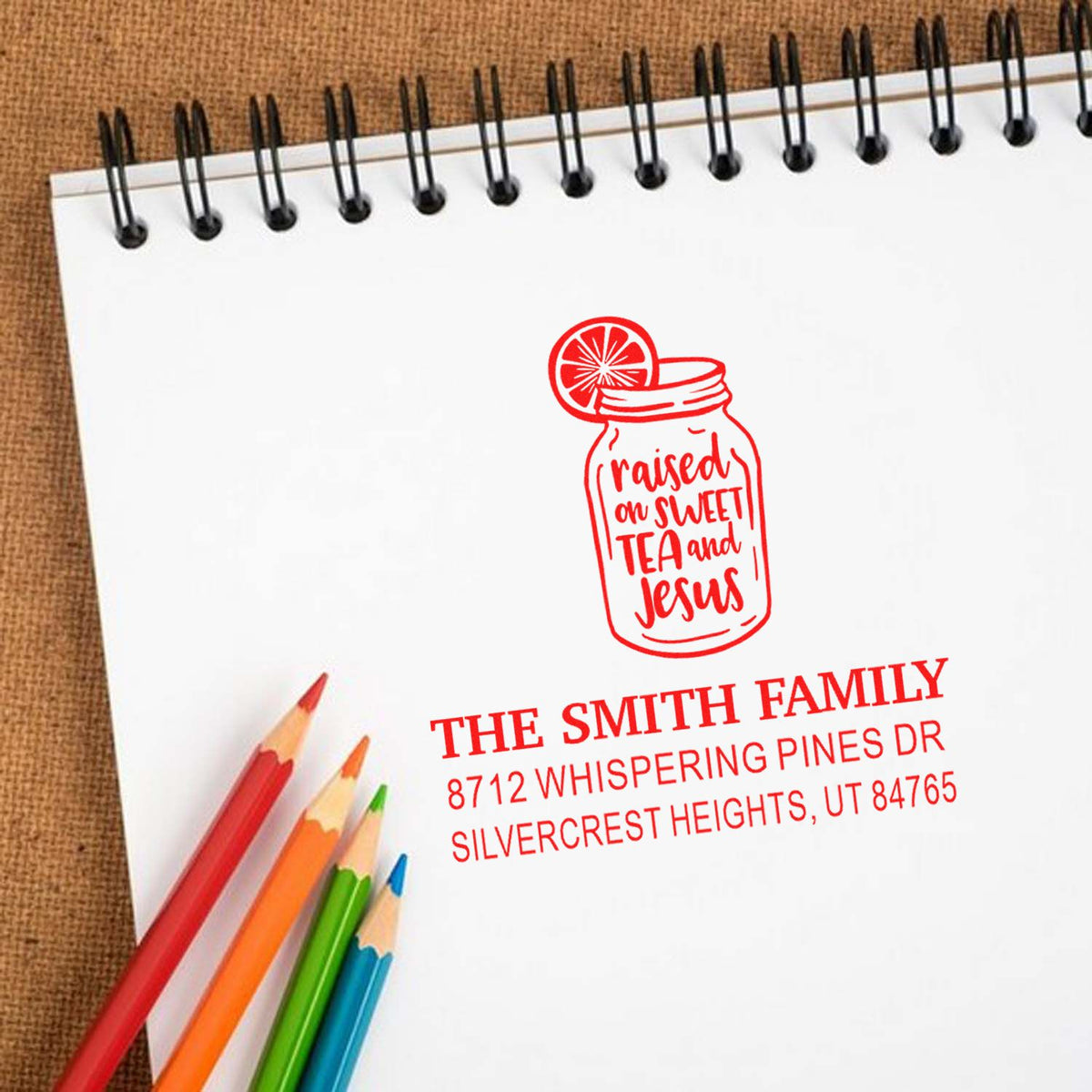 Custom self-inking family stamp on a spiral notebook, featuring a red mason jar raised on sweet tea and Jesus design with colored pencils nearby.